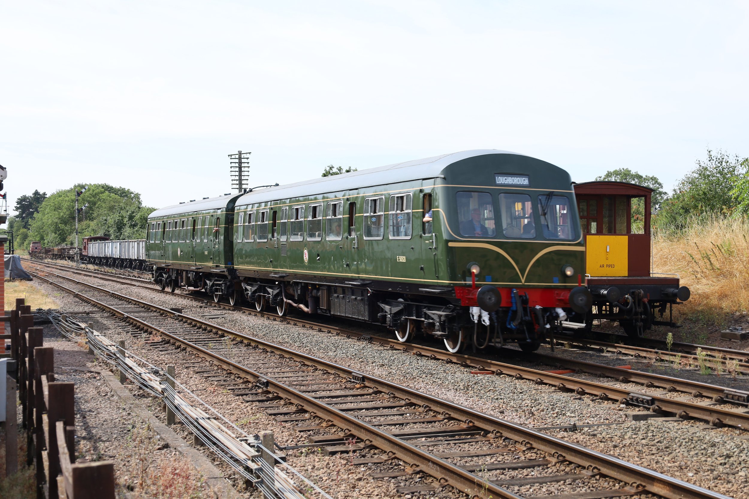 A vintage green train traveling on railway tracks with a sign displaying 'Loughborough,' passing through a rural landscape with trees and a small red and yellow freight shed nearby.