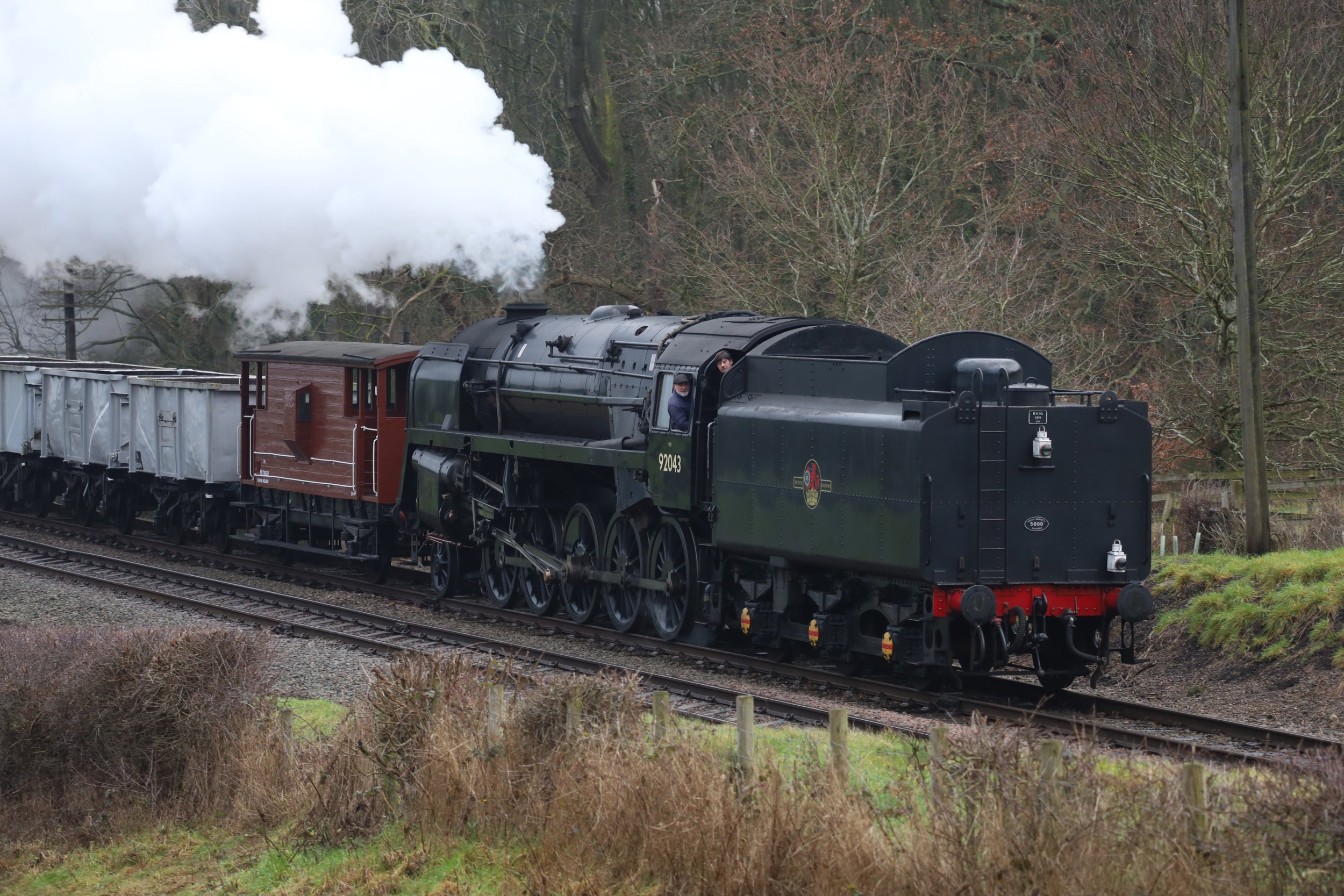 A vintage black steam train with a red and brown wooden carriage moving along a railway track through a wooded area with leafless trees and bushes.