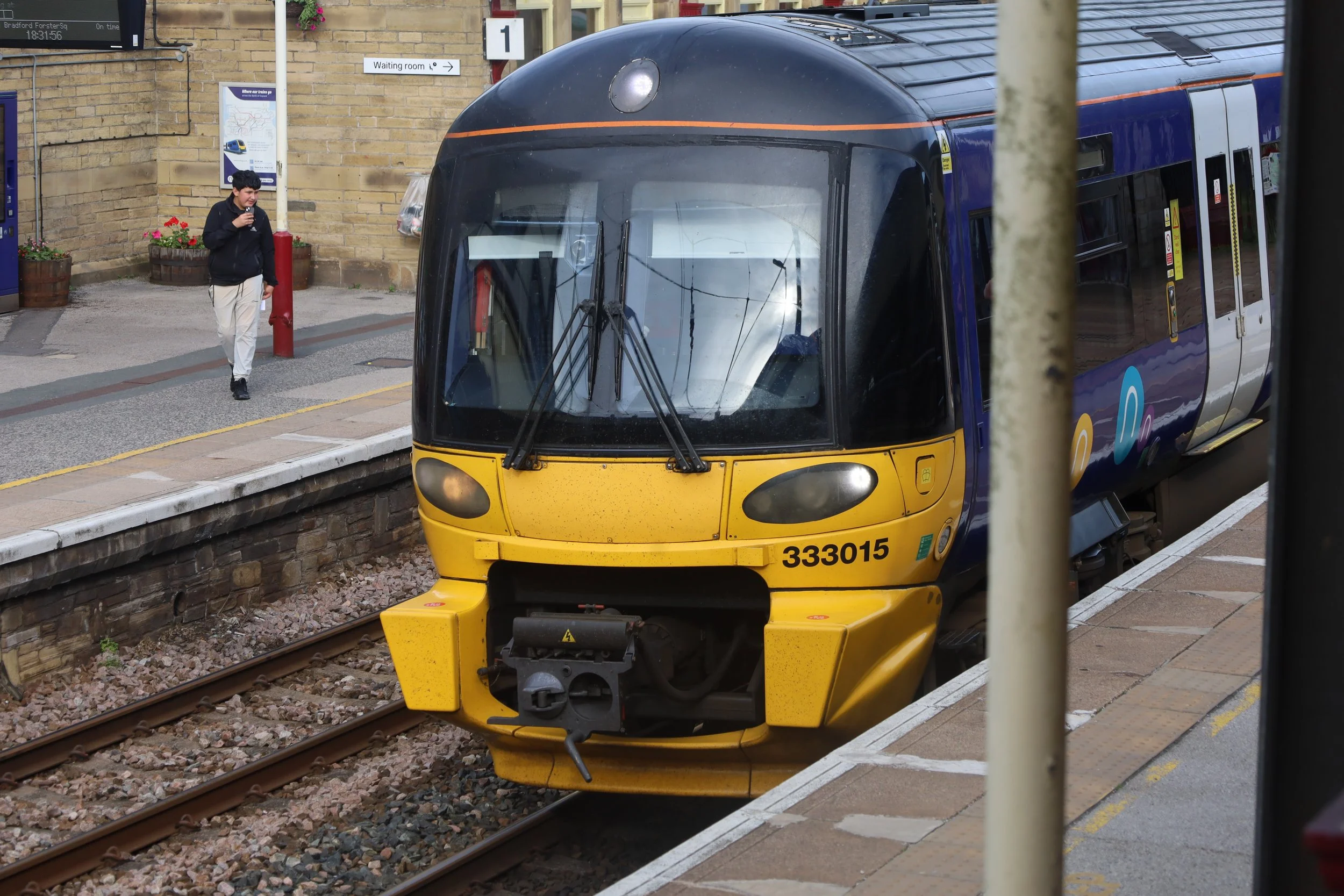 A yellow and blue train at a train station platform with a man walking nearby, and signs indicating a waiting room and train schedule in the background.