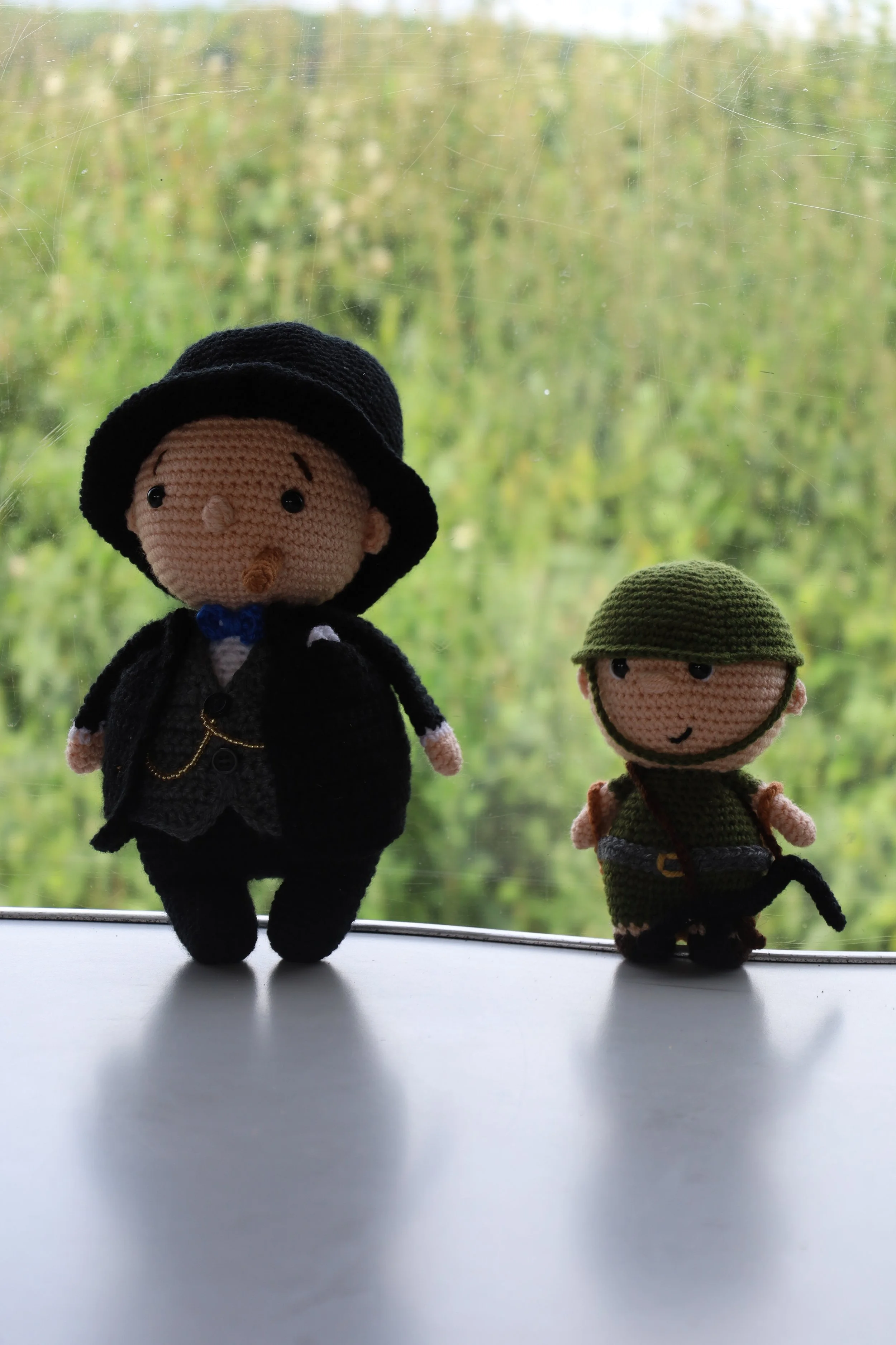 Two knitted dolls dressed as a vampire and a thief stand on a windowsill with a green outdoor background.