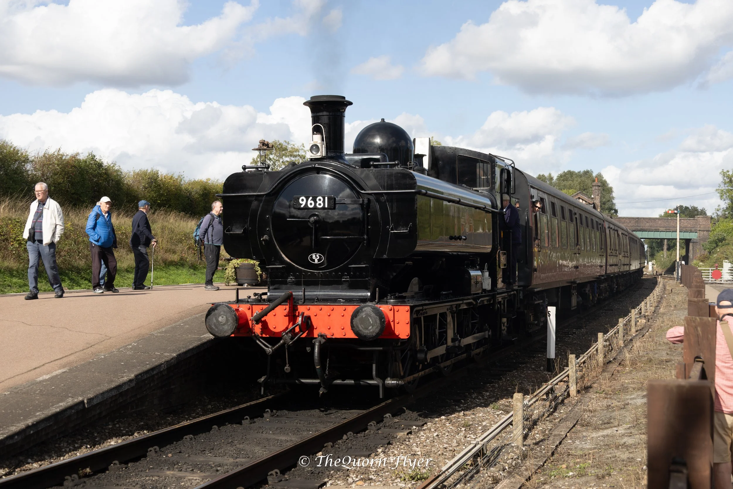 A vintage black steam locomotive train with the number 9681 on the front, pulling along a railway track with people gathered nearby, under a partly cloudy sky.