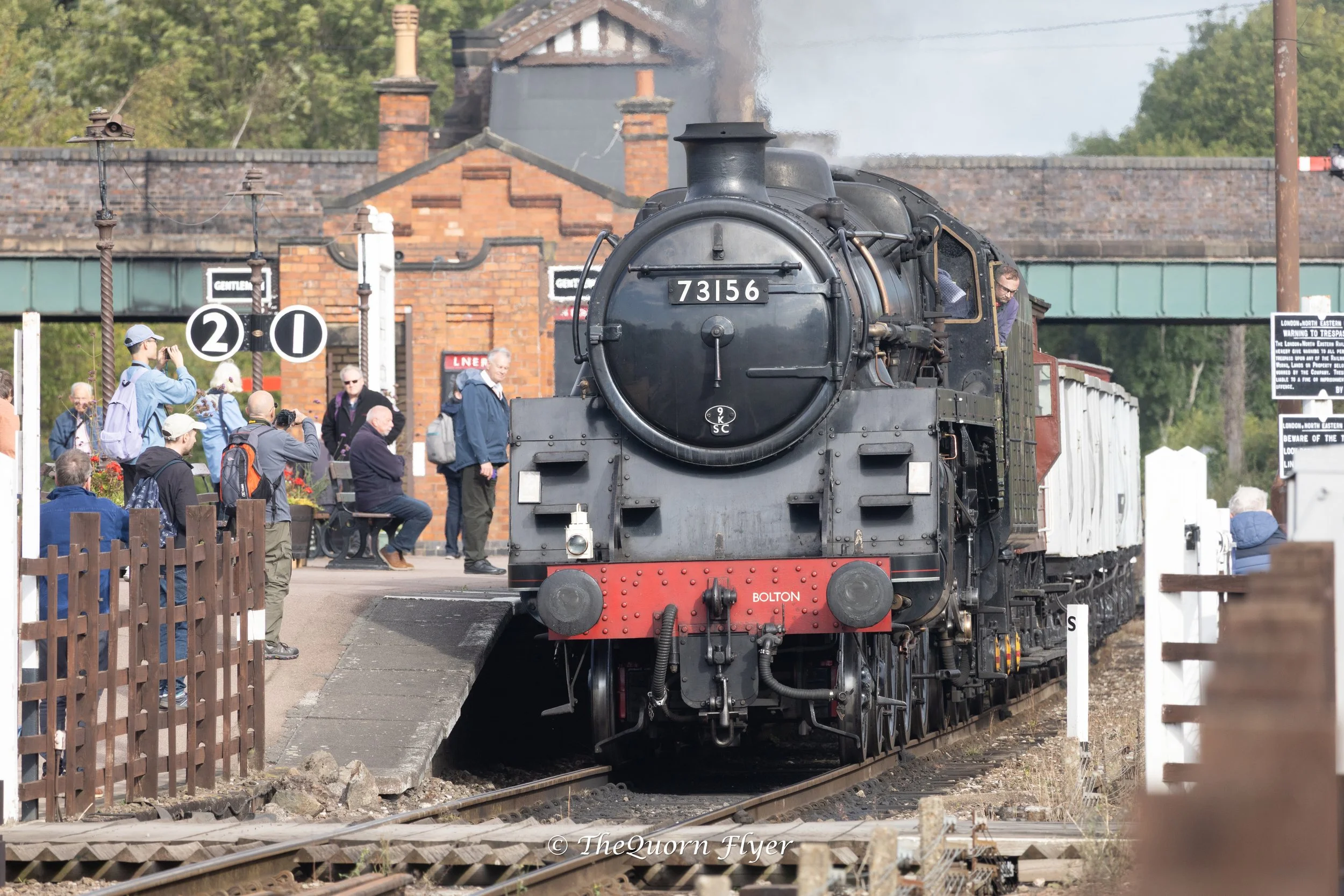 A vintage black steam locomotive with the number 73156 on the front, pulling white passenger cars, arriving at a station platform filled with people taking photos and observing. There are signal signs and a brick station building in the background.