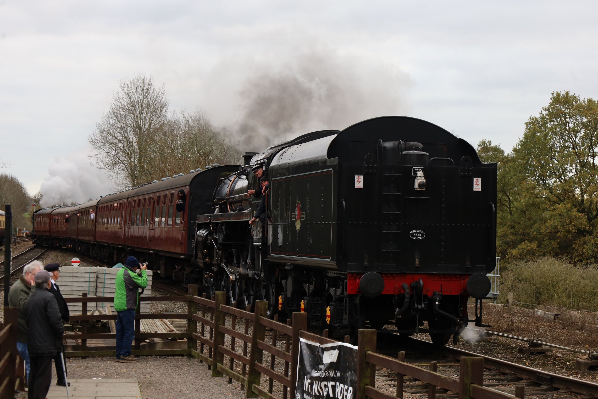 A vintage steam train with black and maroon cars moving on the tracks, people watching and taking photos nearby, and trees with autumn leaves in the background.
