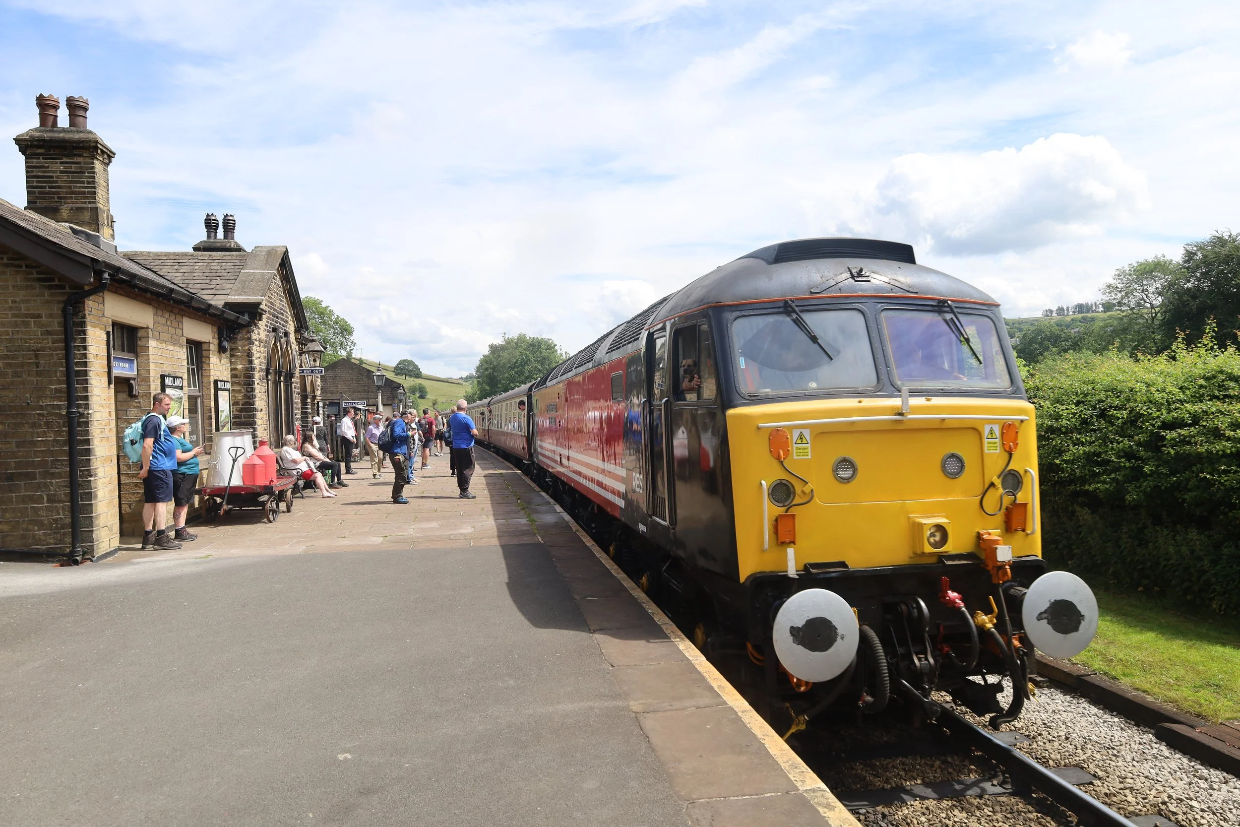 A vintage train at a rural train station with people waiting on the platform and the countryside in the background.