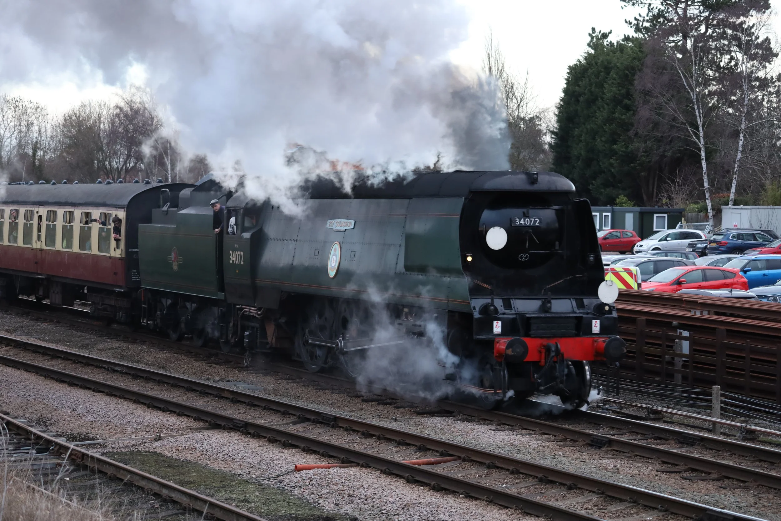 Steam locomotive with smoke and steam emitting from its stack, traveling on railway tracks in a parking lot with parked cars and some trees in the background.