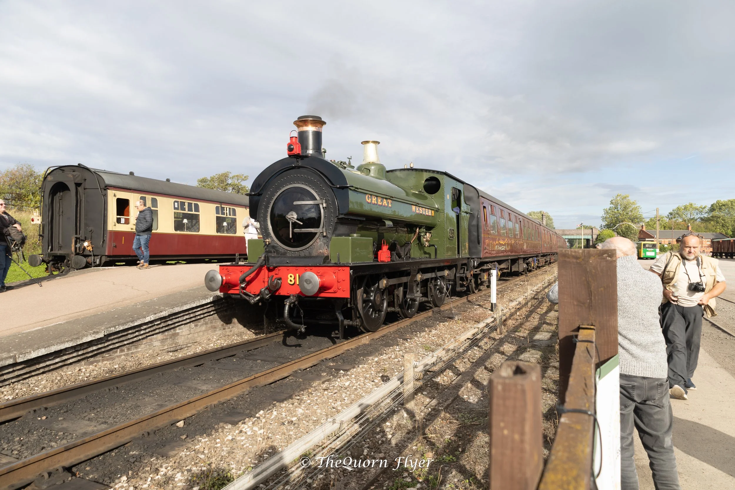 A vintage green steam train with red accents labeled 'Great Western' at a train station platform, with several people nearby, some taking pictures.