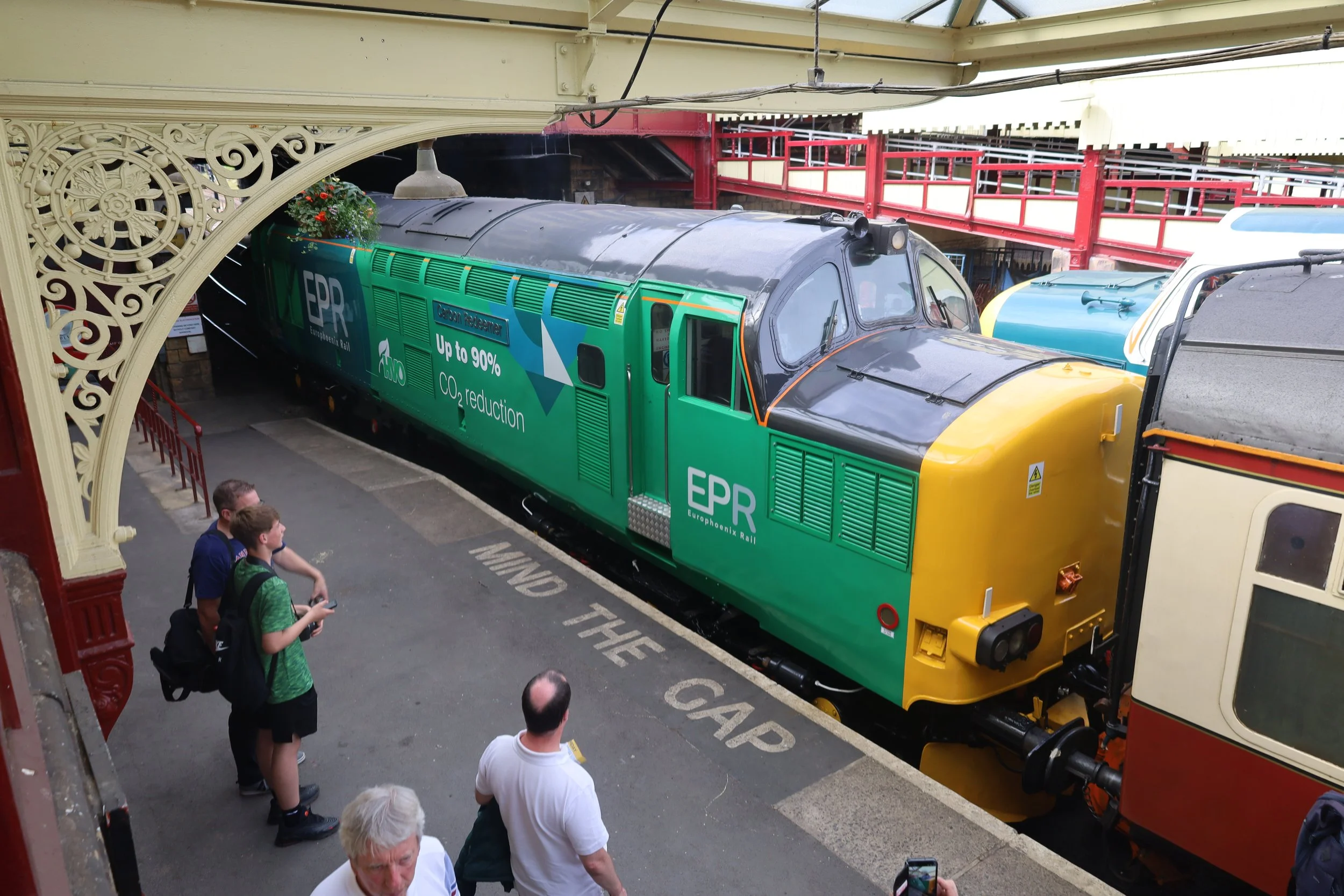 A colorful train with a green and yellow engine, parked underneath a decorative iron archway, with several people standing nearby taking photos and looking at the train.