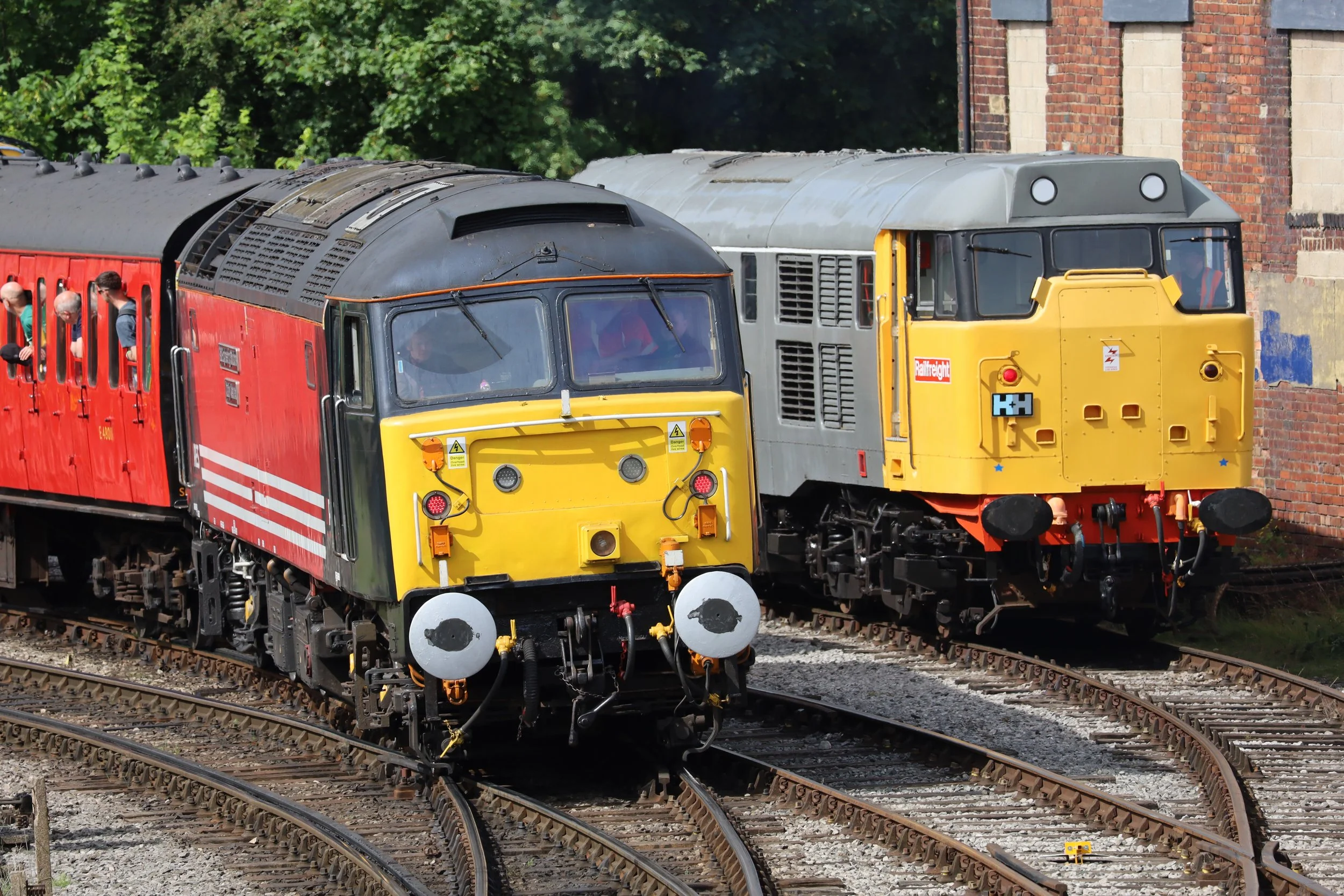 Two railway locomotives, one yellow and one gray, on railway tracks with a brick building and green trees in the background.