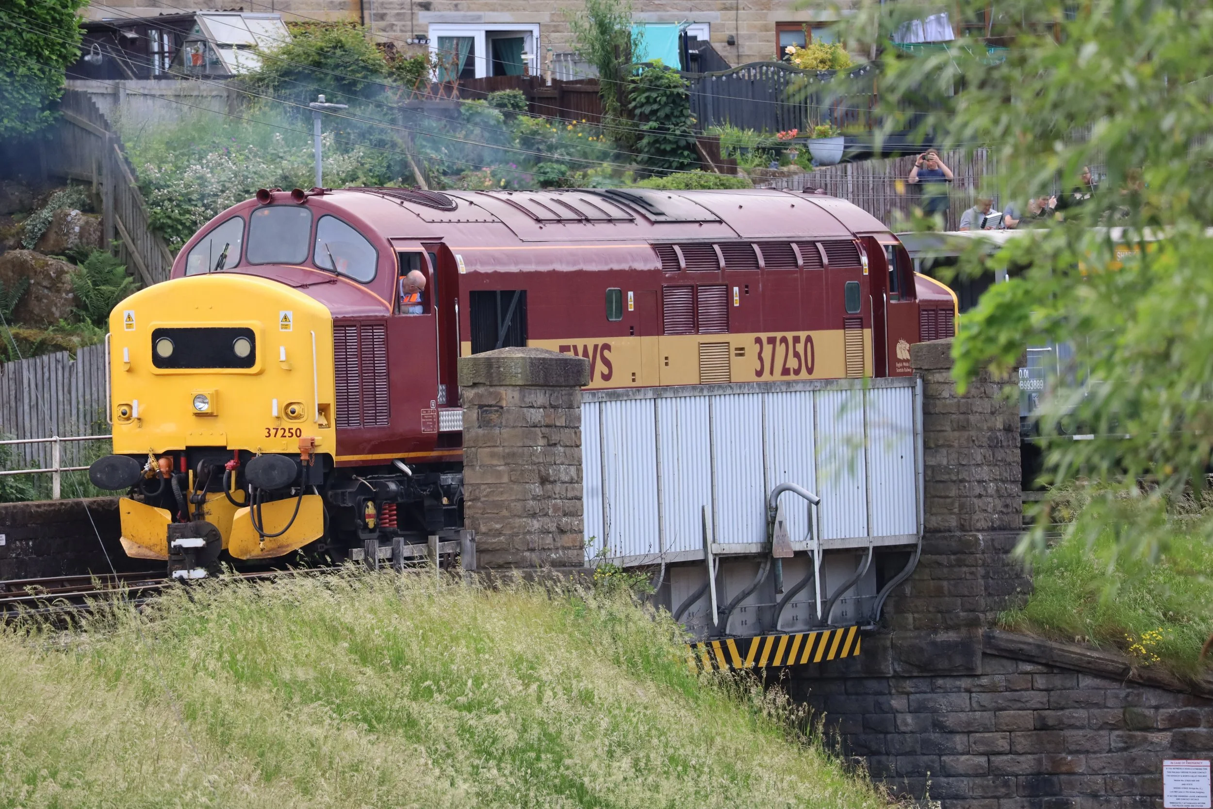A red and yellow train car passing over a stone bridge in a residential area with houses and gardens. Some people are taking photographs of the train from behind a fence.