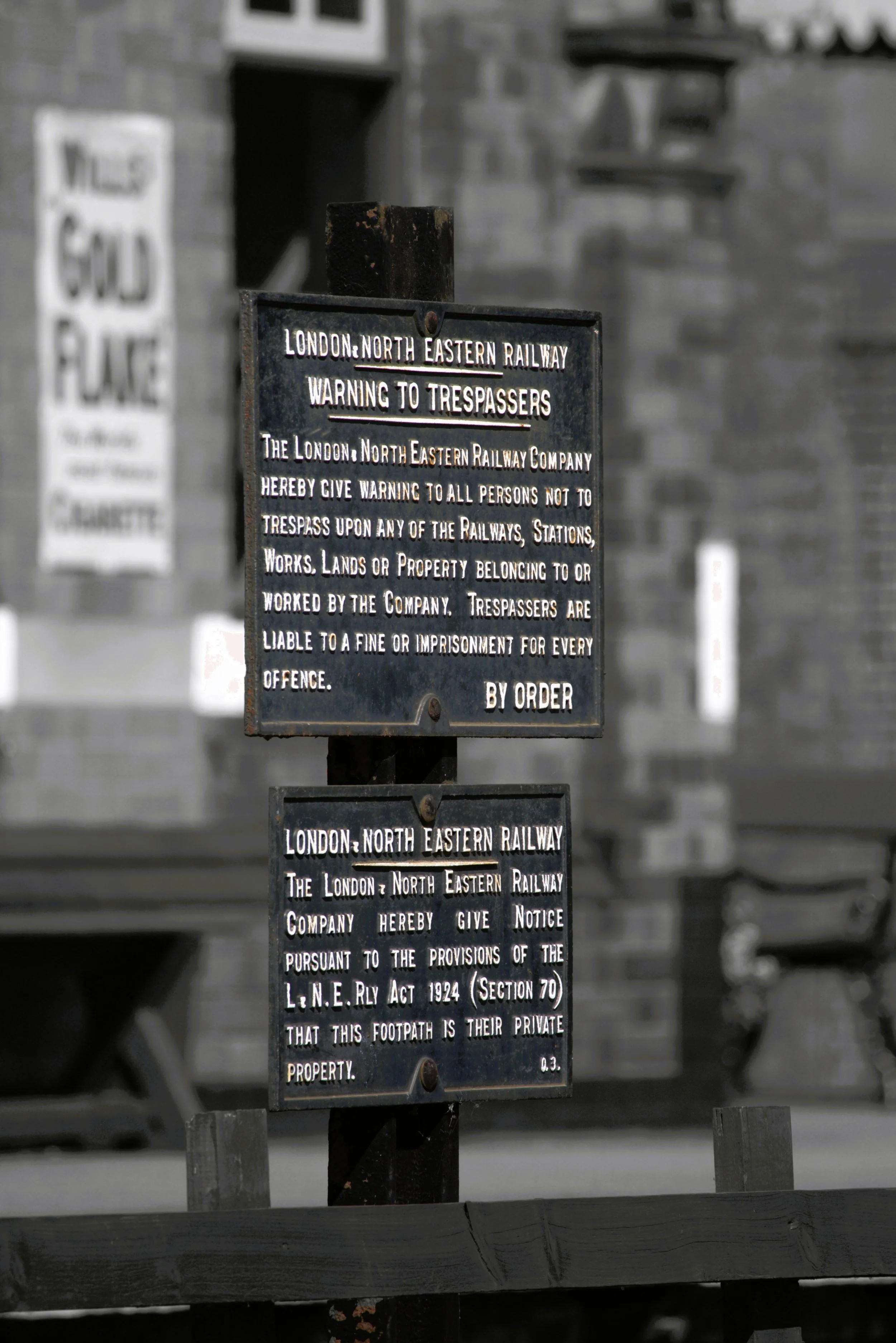 A black metal sign with white engraved text warning trespassers not to enter private railway property, and detailing legal consequences of trespassing. In the background, there is another similar sign and a brick wall.