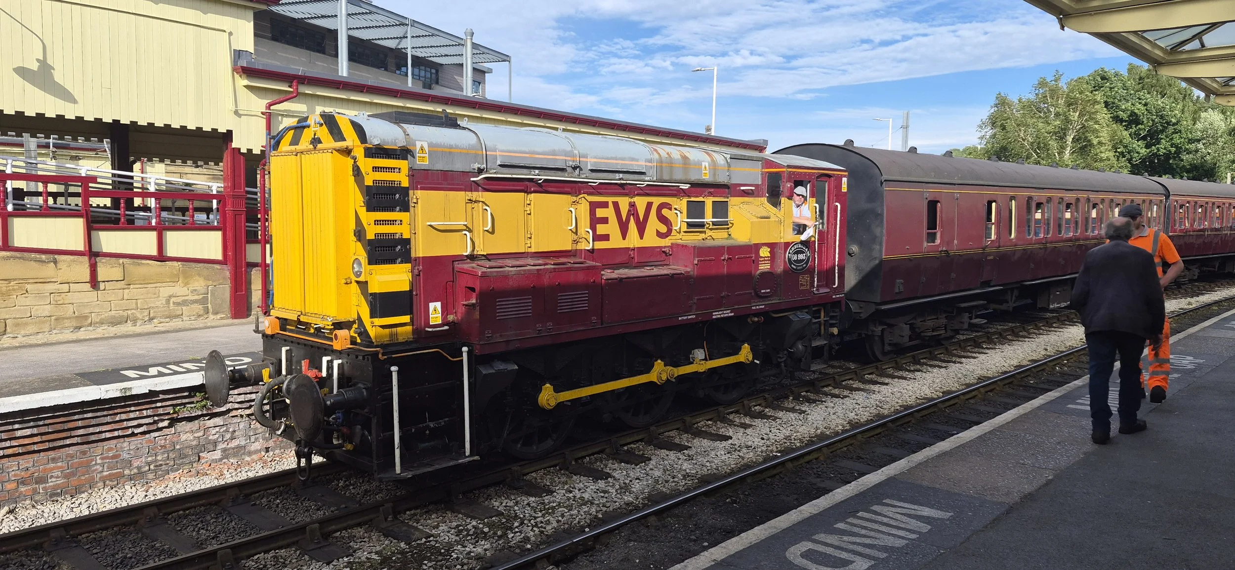 A vintage train at a station with a yellow and red engine marked 'EWS' and a maroon passenger carriage. People are standing near the train, and the station platform is visible.