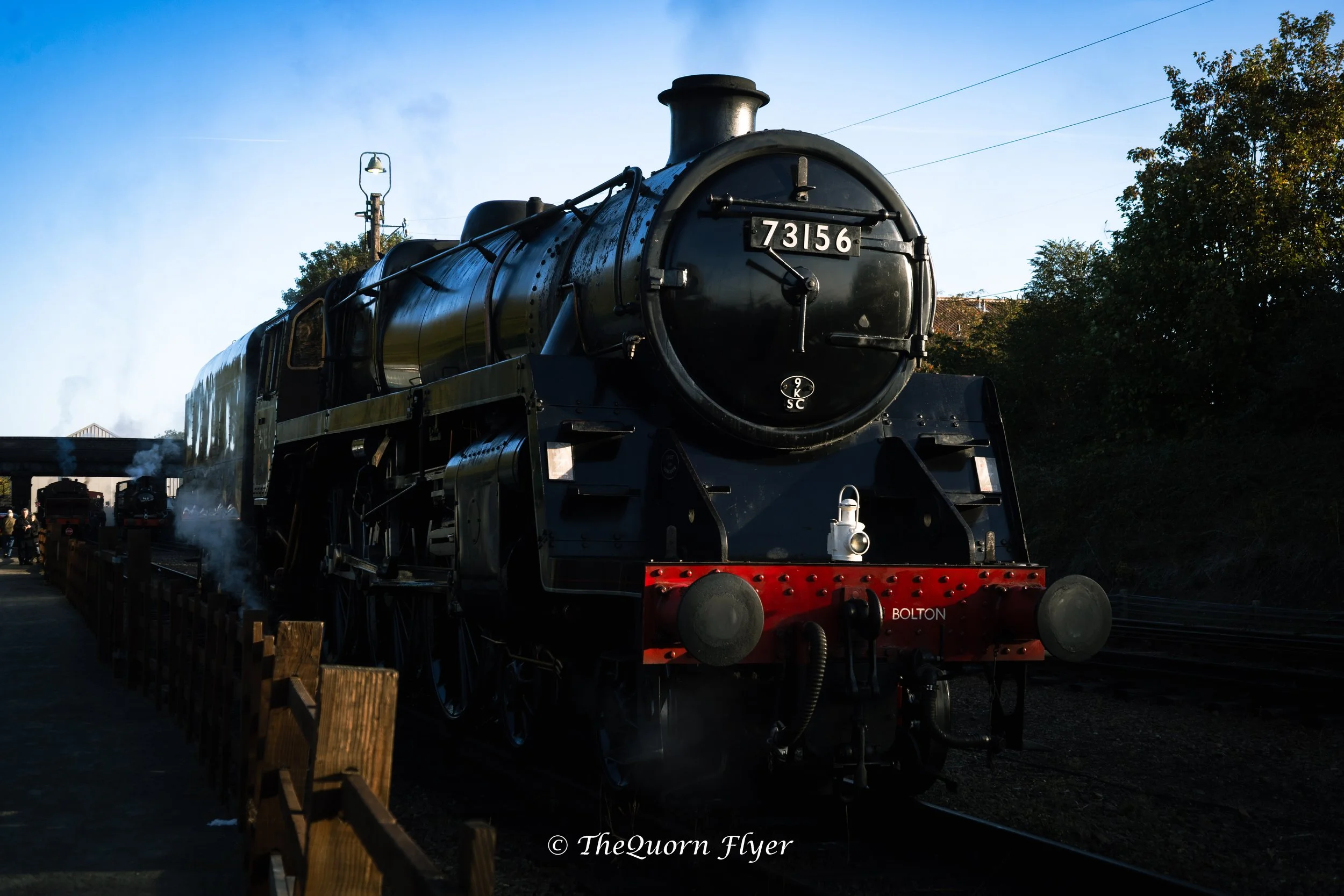 A black steam train with the number 73156 on the front is on a railway track outdoors. There are trees on the right side and a clear blue sky above.