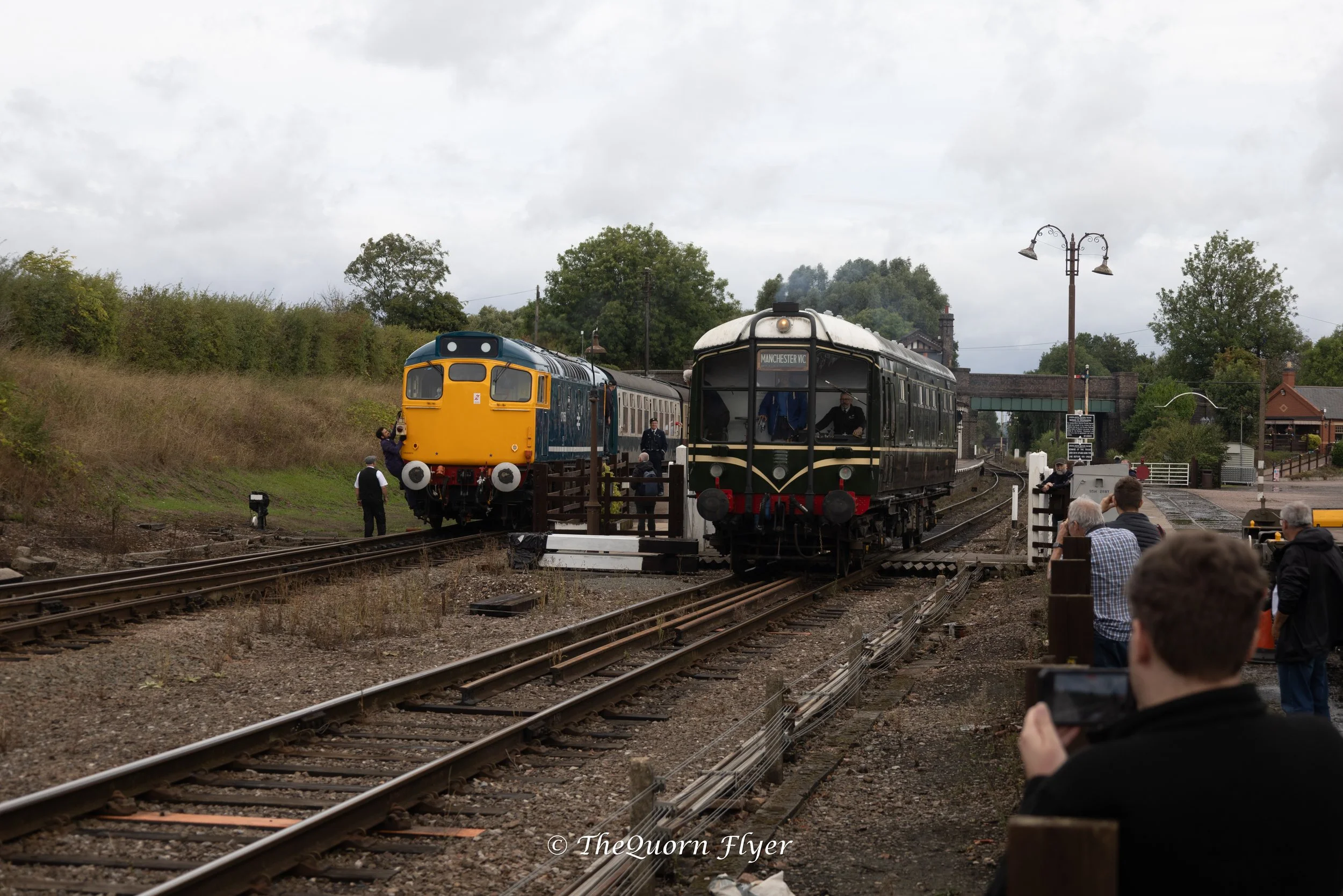Two vintage trains, one yellow and blue, and one black with cream accents, are positioned on parallel train tracks at a railway station. Several people are gathered around, some taking photos. The station is outdoors with trees, a building, and a cloudy sky in the background.