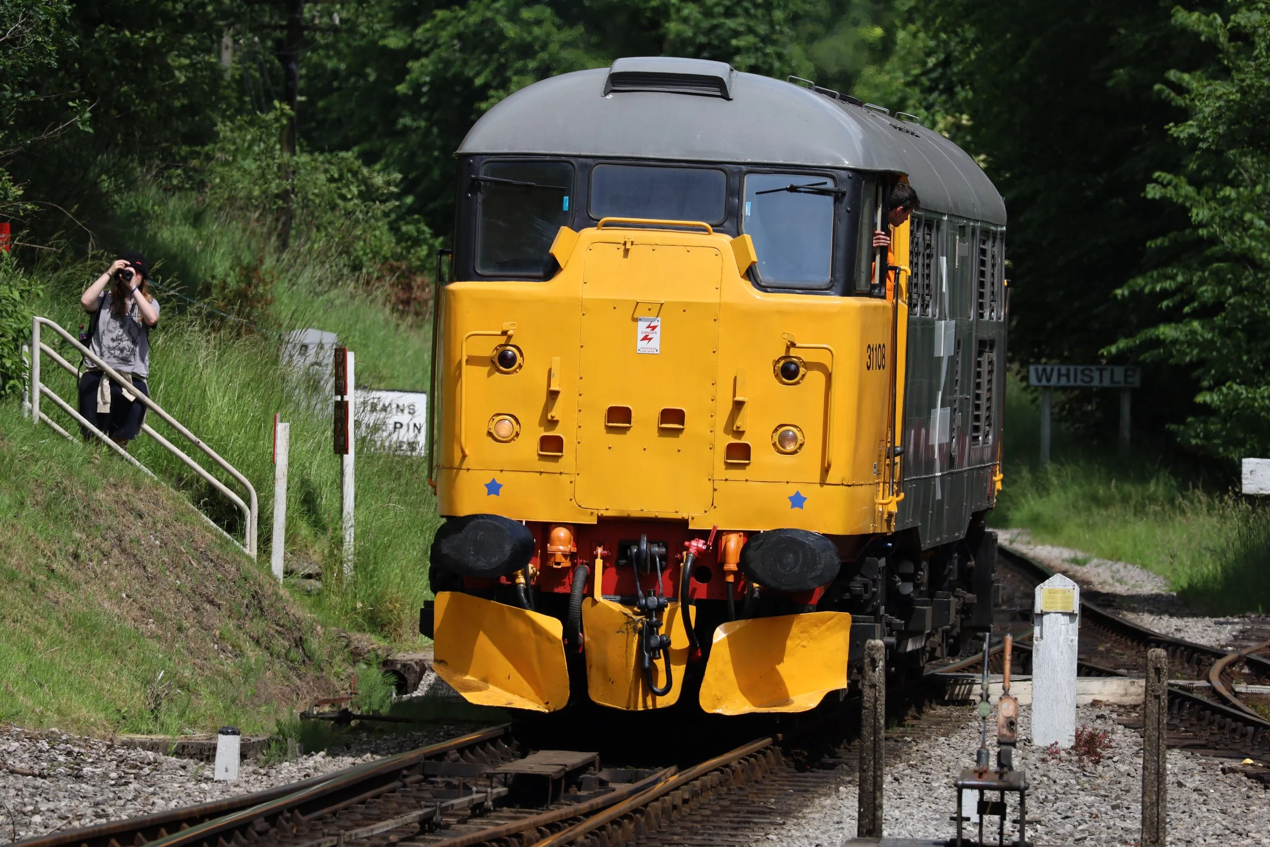 A yellow and gray vintage locomotive train on railway tracks with a person taking a photo nearby, surrounded by green trees.