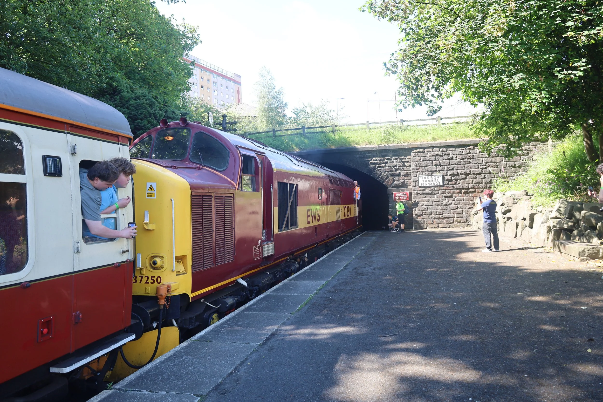 A vintage red and yellow train stopped at a platform under a stone tunnel with a sign indicating it is a 150-yard long tunnel. There are people around the train, some taking pictures and some waiting. The background includes green trees, a grassy hill, and a tall building.