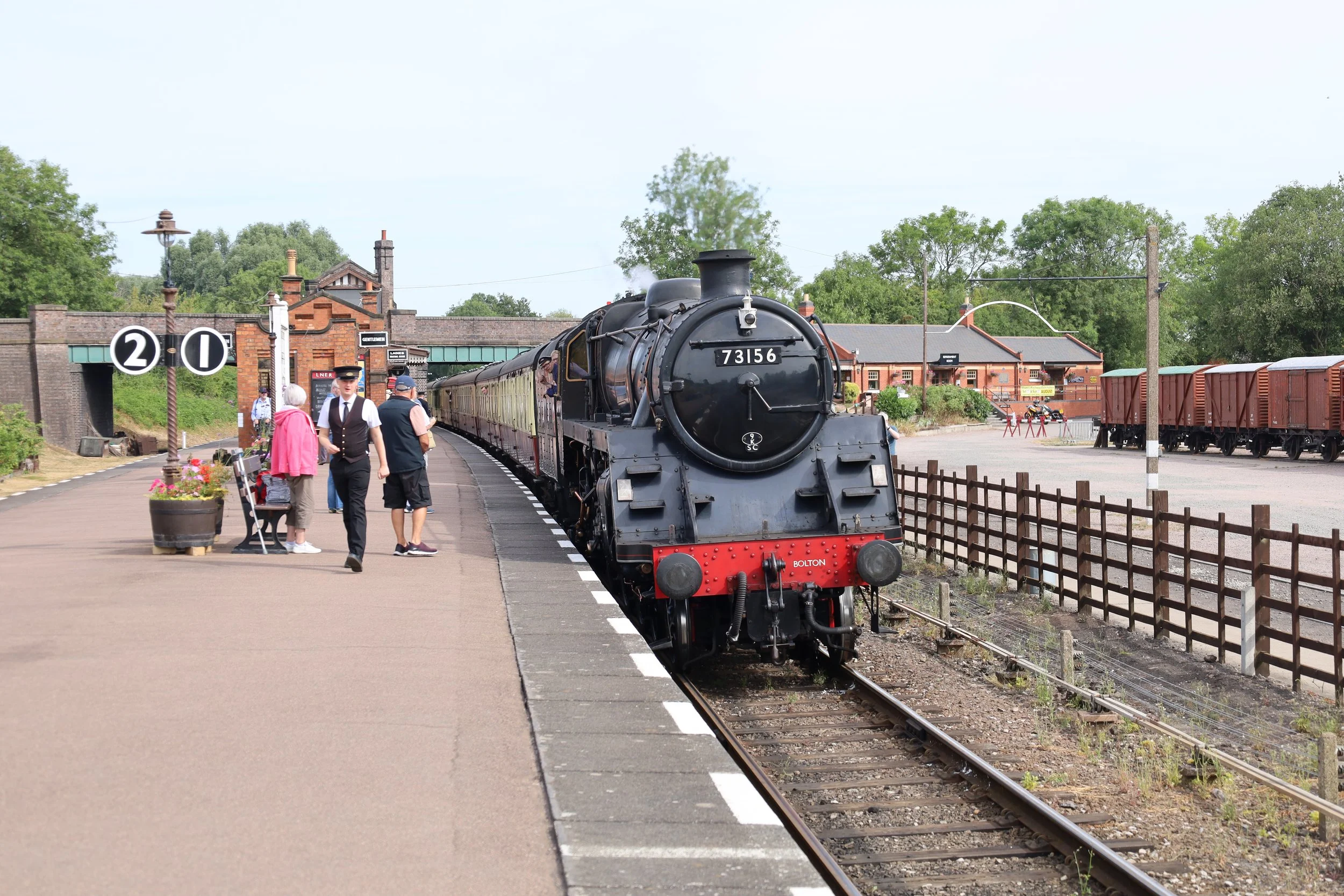 A vintage steam train at a railway station platform with people, including a conductor, standing nearby. The train is black with the number 73156 on the front, and there are red freight cars on the track. The station building and trees are visible in the background.