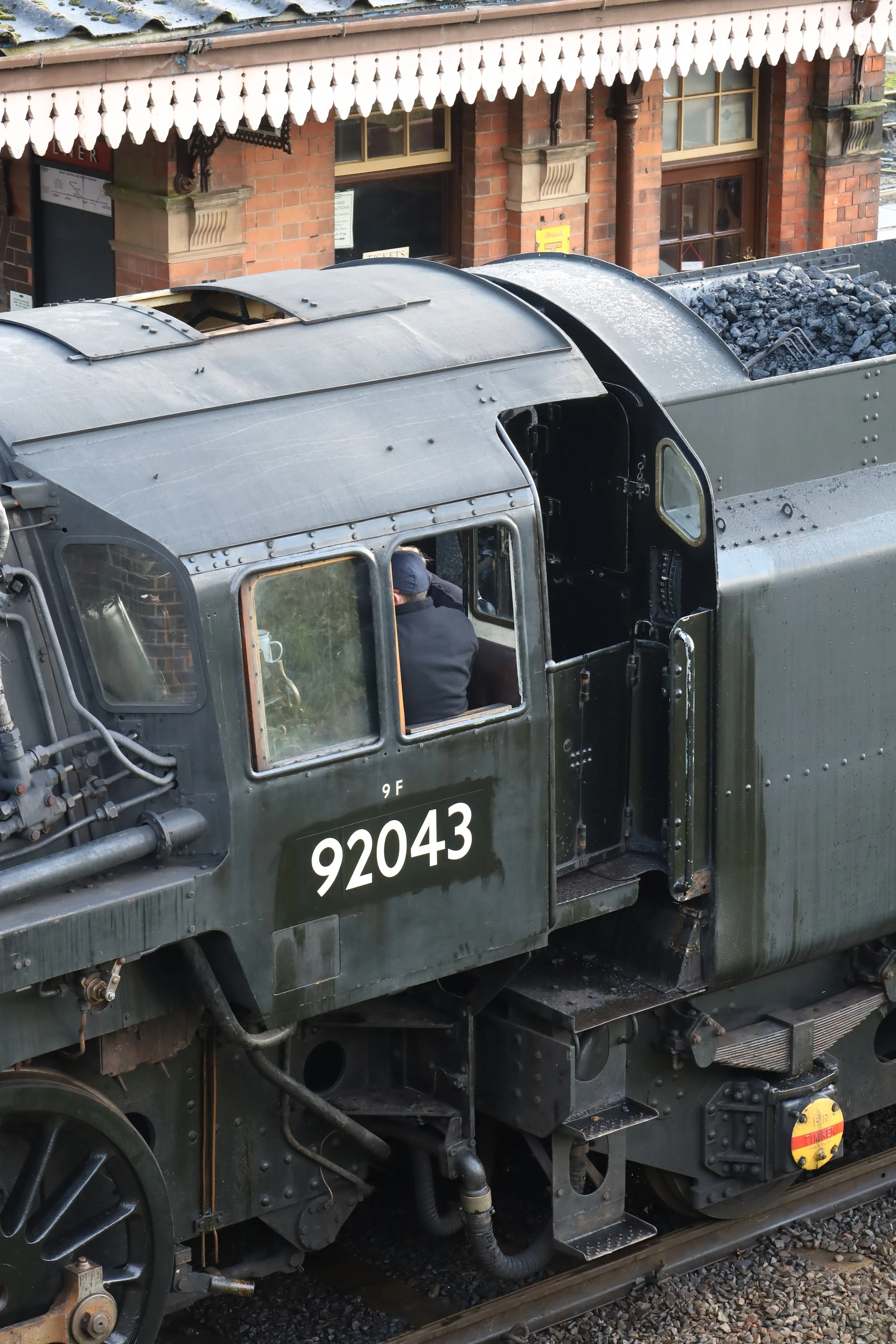 A vintage steam locomotive with the identification number 92043, parked on the railway tracks next to a brick building with decorative elements and windows.