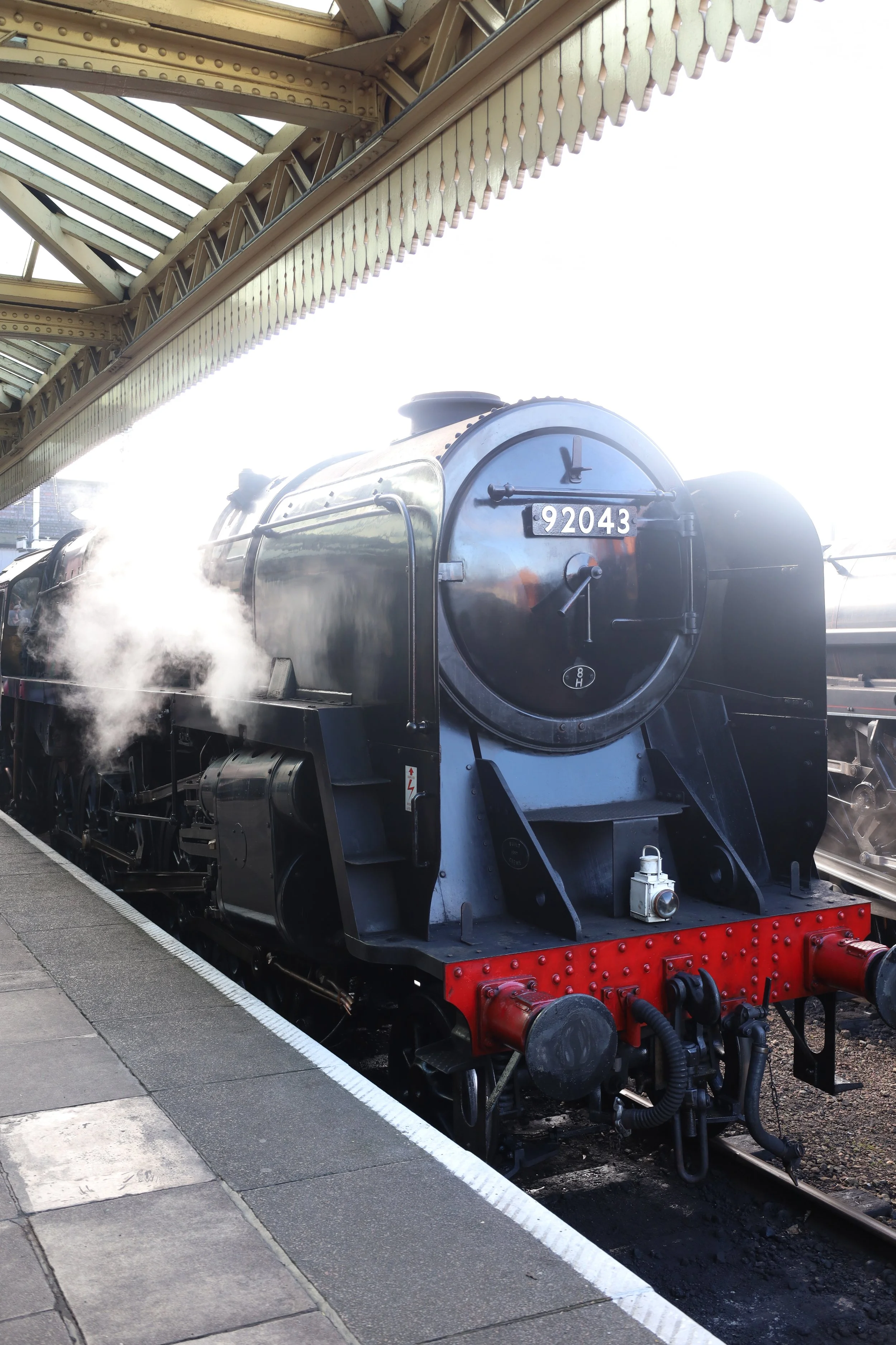 A black steam train with the number 92043 on the front, emitting steam, parked at a station platform beneath a metal canopy with decorative trim.