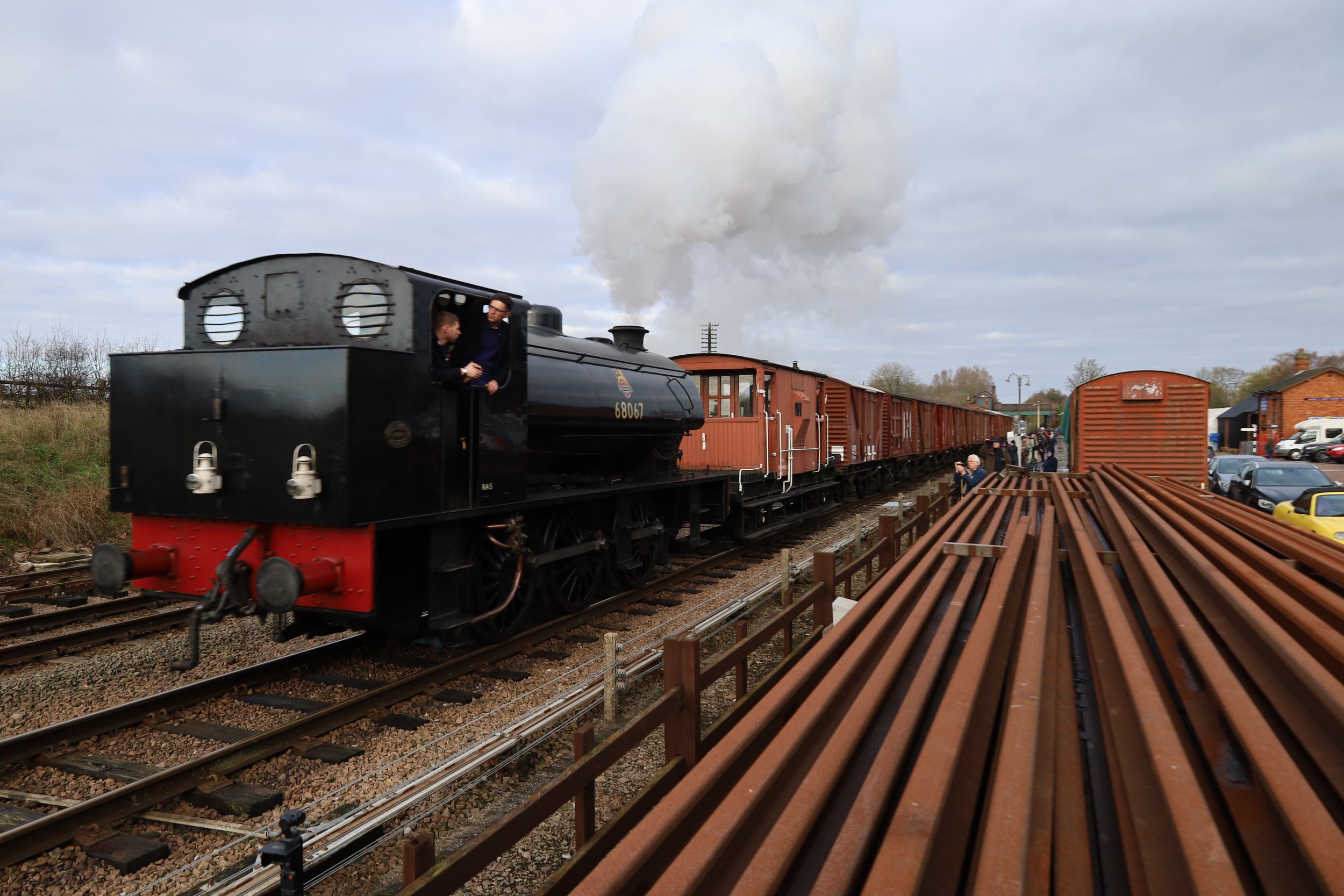 A black steam train pulling brown freight cars on railway tracks with people observing nearby, under a cloudy sky.