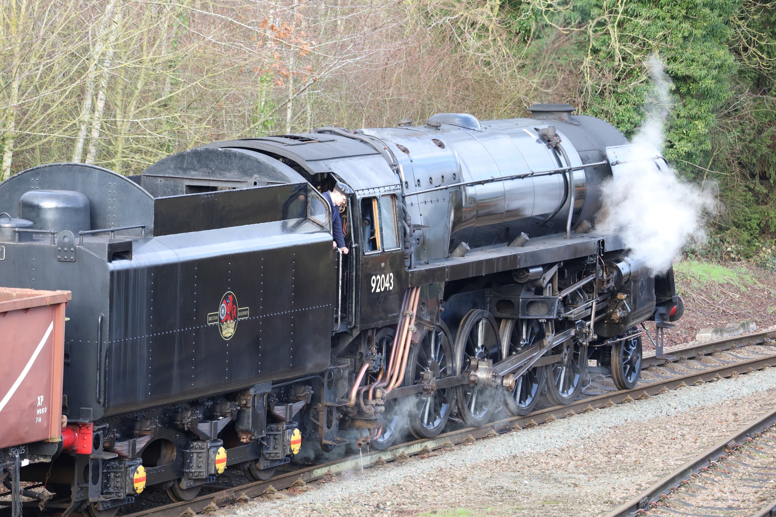 Black steam locomotive moving on railway tracks with steam coming from the front, surrounded by trees and greenery.