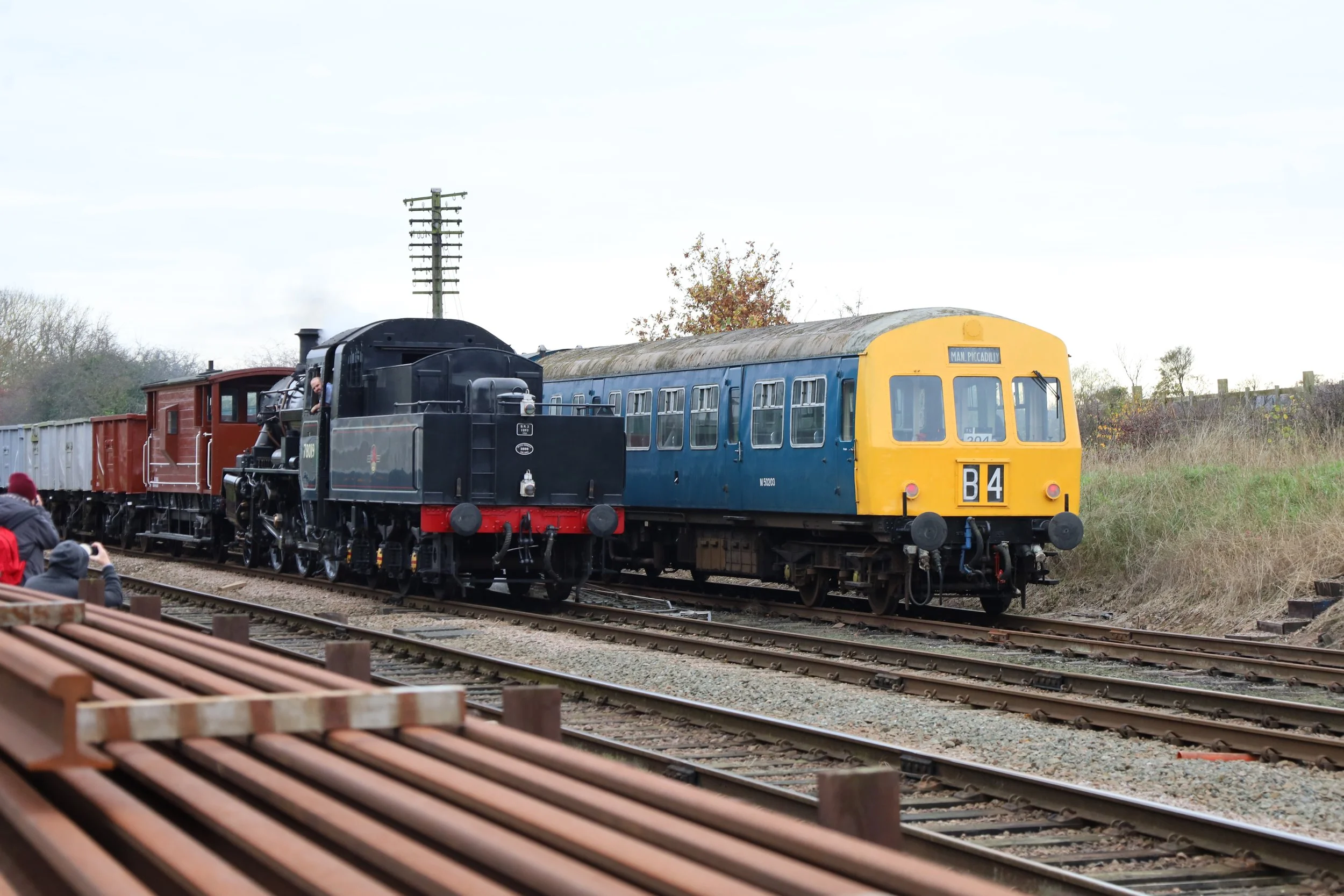 A vintage steam train moving along a railway track during daytime, with a person visible in the engine cab. The train consists of a black engine, a blue passenger car with a yellow front, and freight cars behind. There are spectators, some taking photos, on the side of the tracks, and trees and utility poles are visible in the background.
