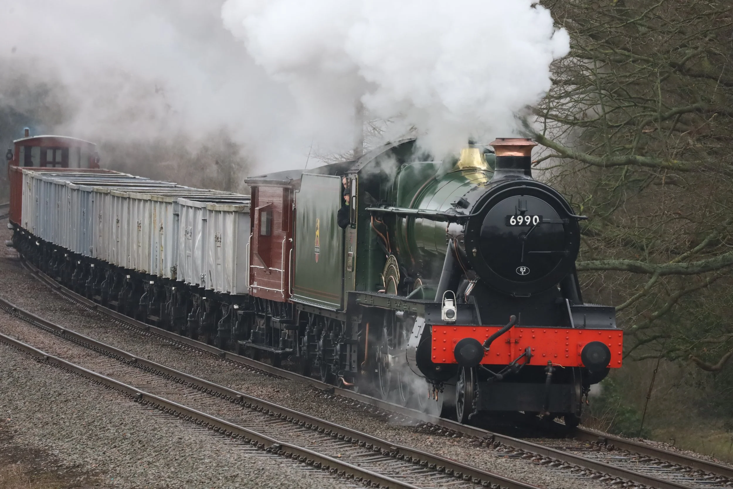 A vintage steam train with green and black colors and the number 6990 on the front, emitting white smoke while moving on railway tracks through a wooded area.