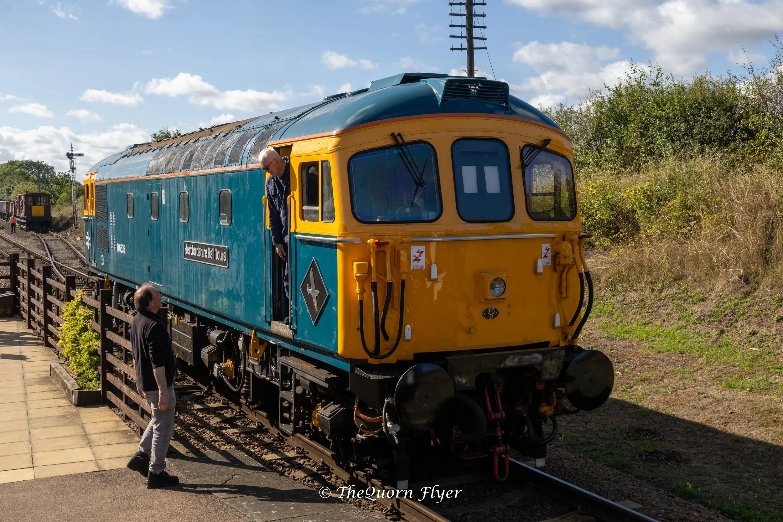 A vintage blue and yellow train carriage on a railway track with a man leaning out of the window and two men standing nearby, under a partly cloudy sky.