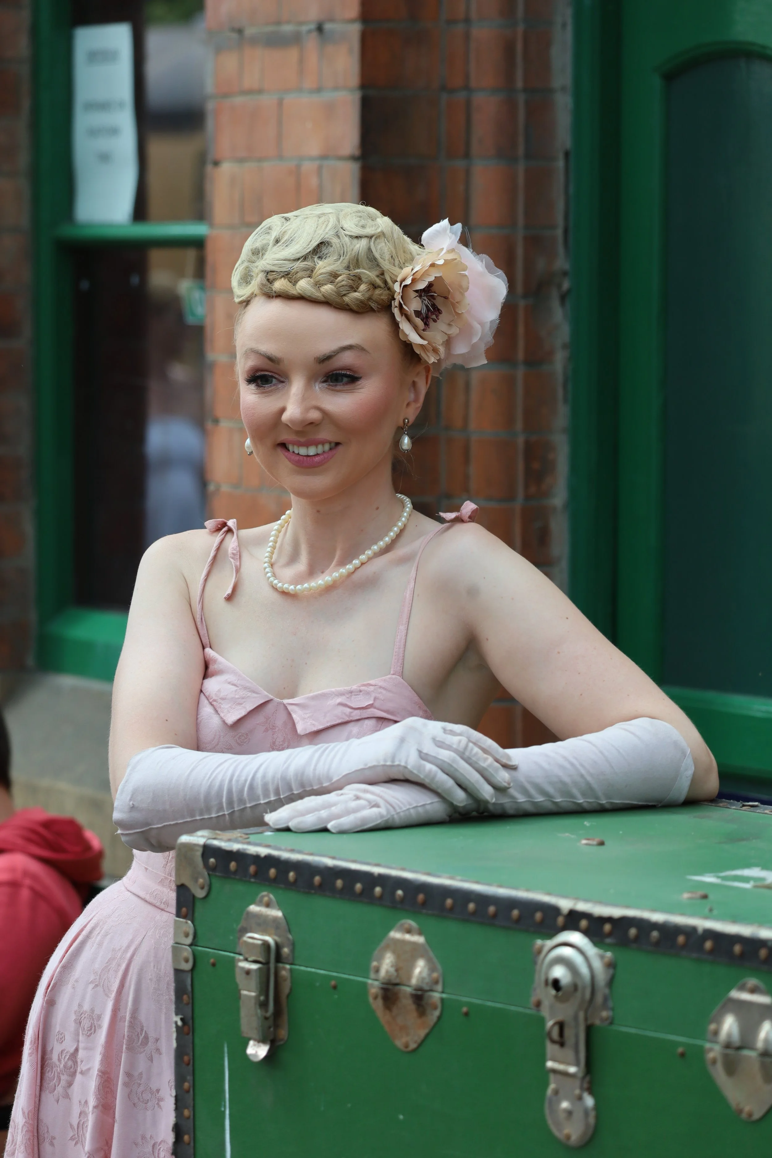A woman dressed in vintage style clothing, including a pink dress, pearl necklace, and long gloves, is smiling while leaning on a green trunk. She has short, curly blonde hair adorned with a large flower accessory. The background features a brick wall and a green window frame.