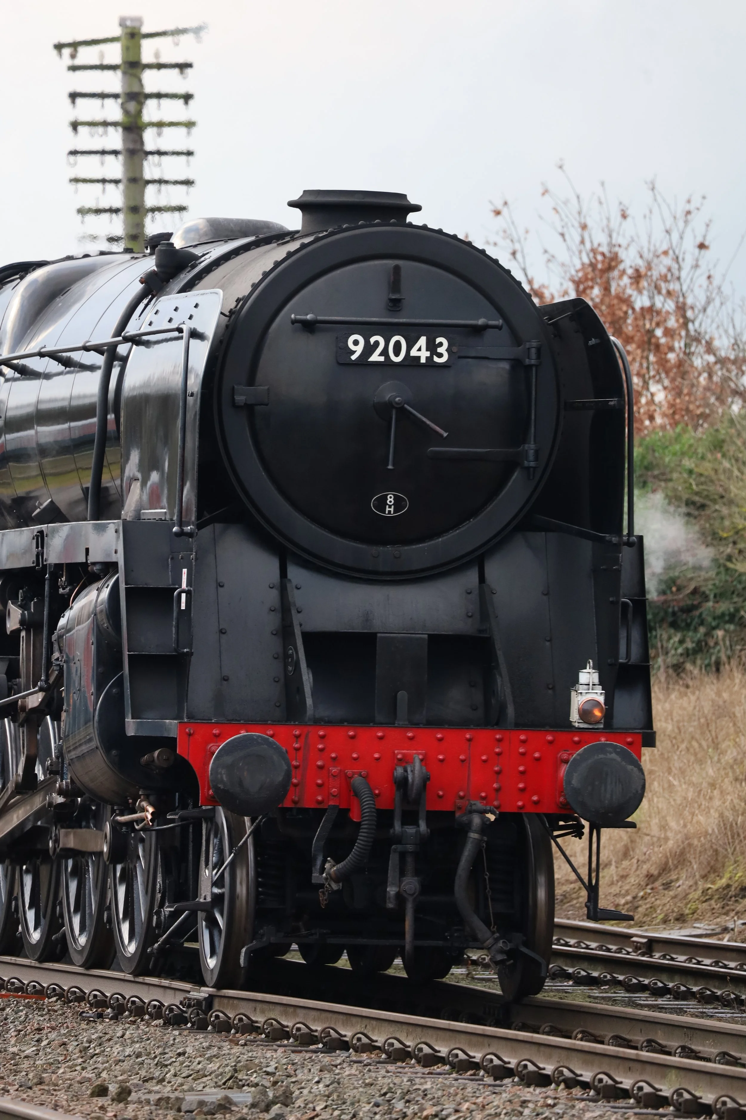 A black vintage steam locomotive with the number 92043 on the front, moving on railway tracks with trees and electricity poles in the background.