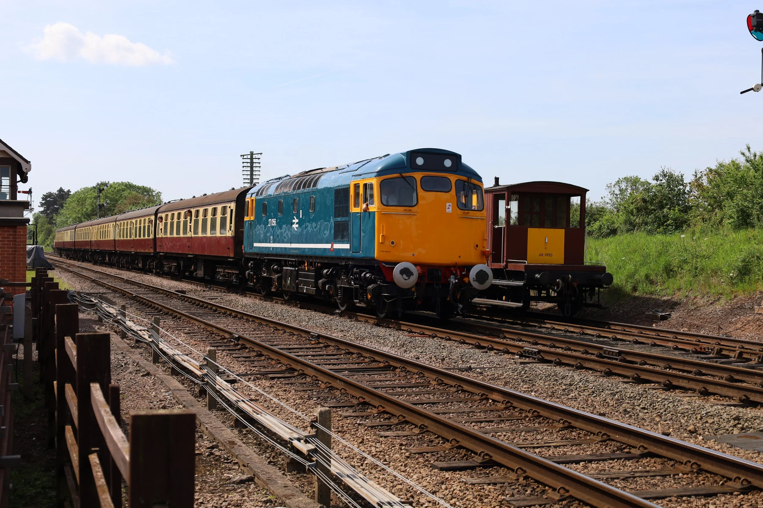 A blue and yellow diesel locomotive pulling yellow and maroon passenger cars along railway tracks in a rural area with green trees and blue sky.