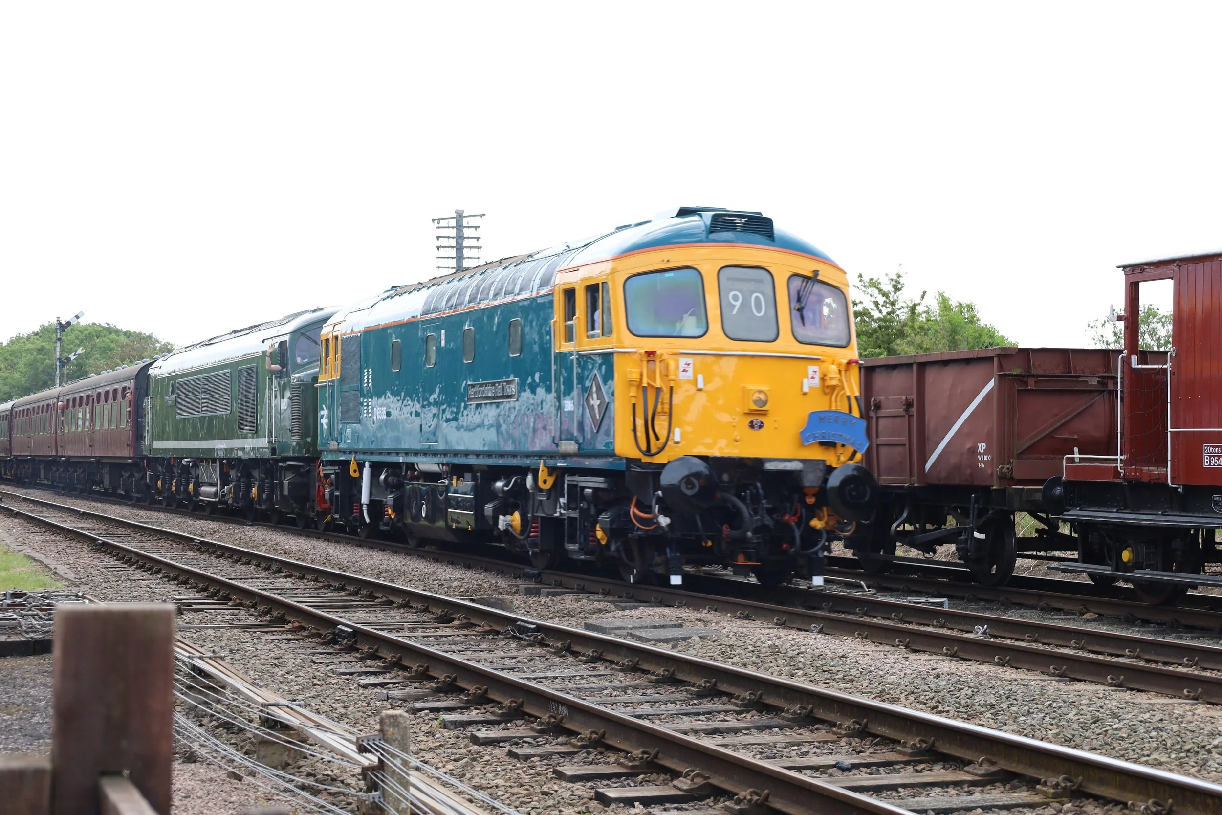 A vintage train with blue and yellow locomotive and red passenger cars on railway tracks, with green trees and utility poles in the background.