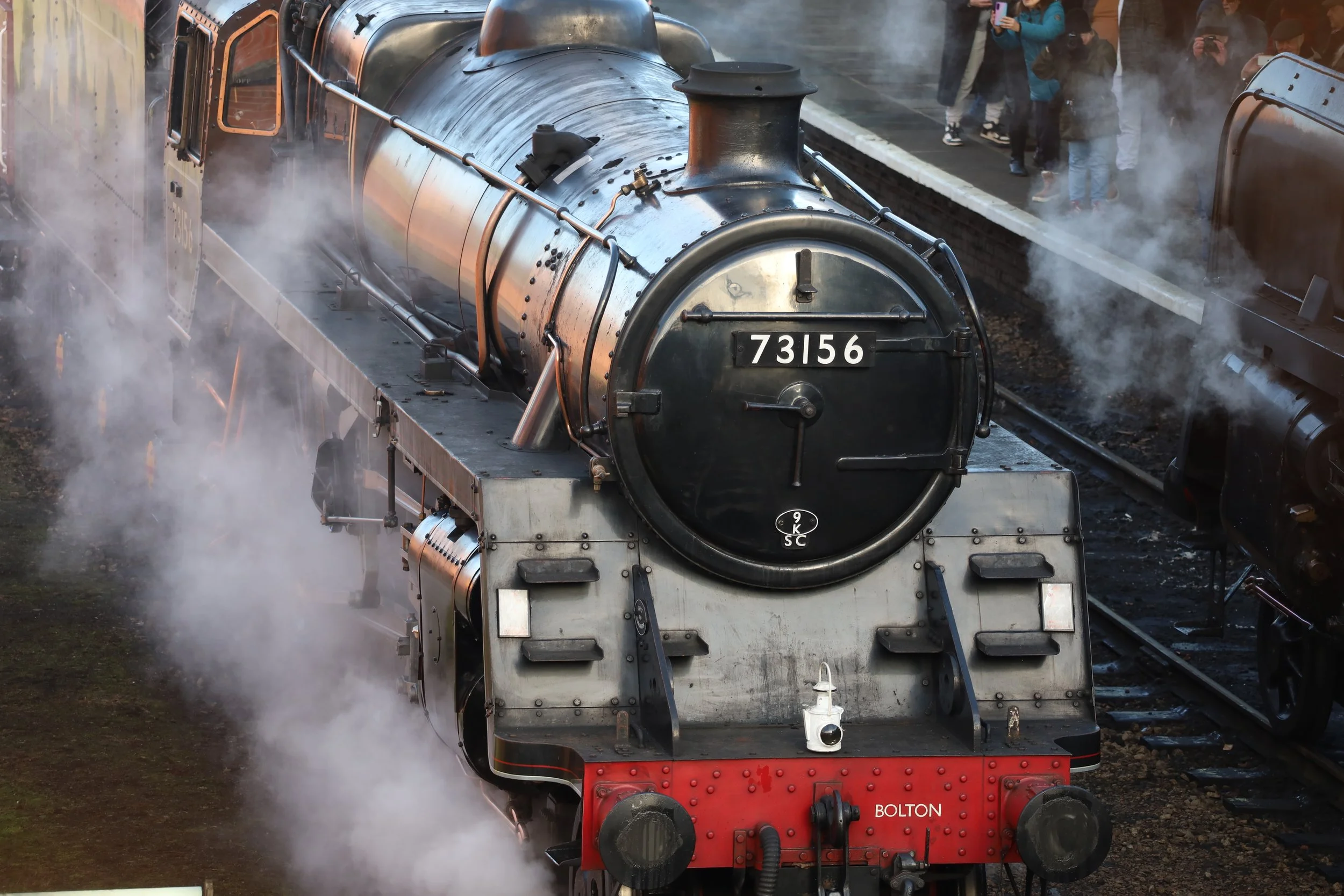 A vintage steam locomotive with the number 73156 on the front, emitting steam and smoke, with spectators taking pictures on a platform in the background.
