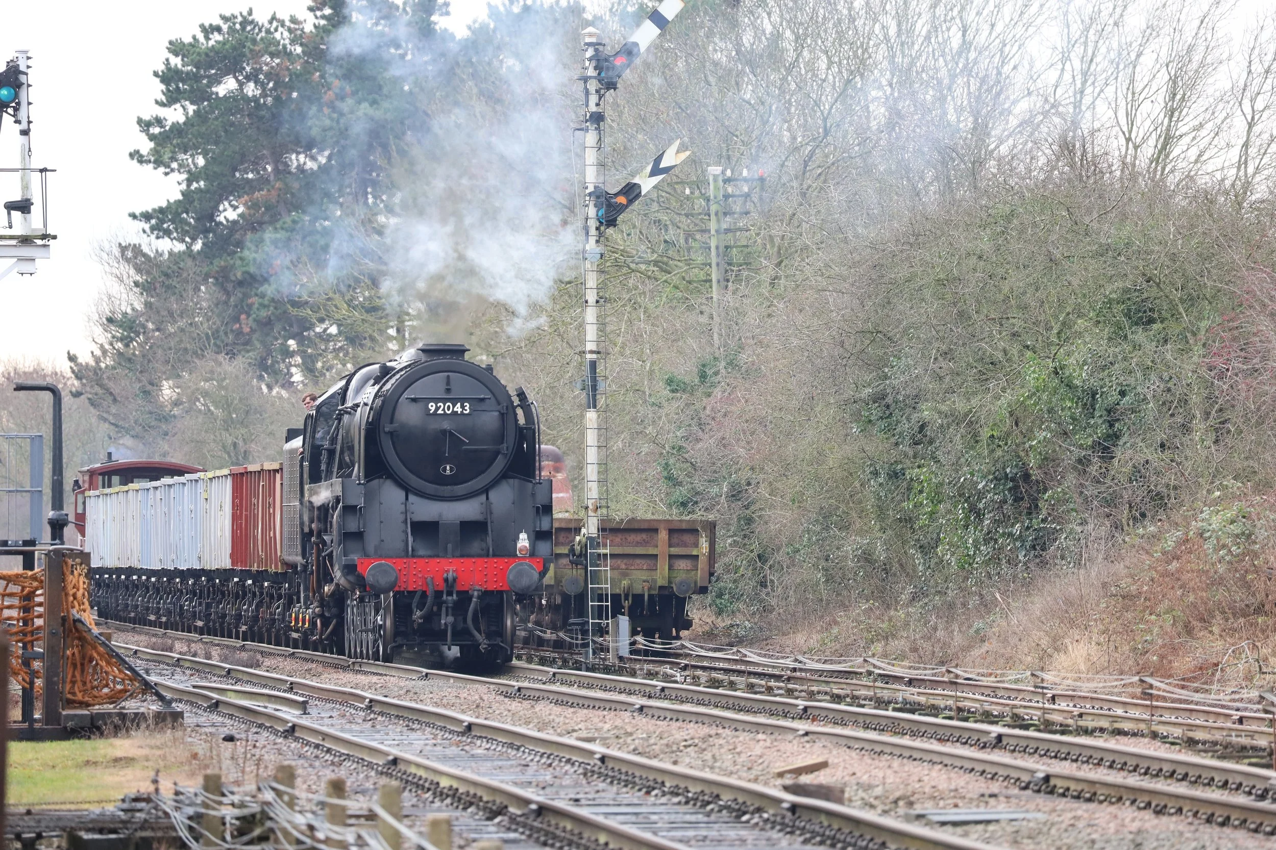 A black steam locomotive train moving on railway tracks through a rural area with trees and signal lights in the background.