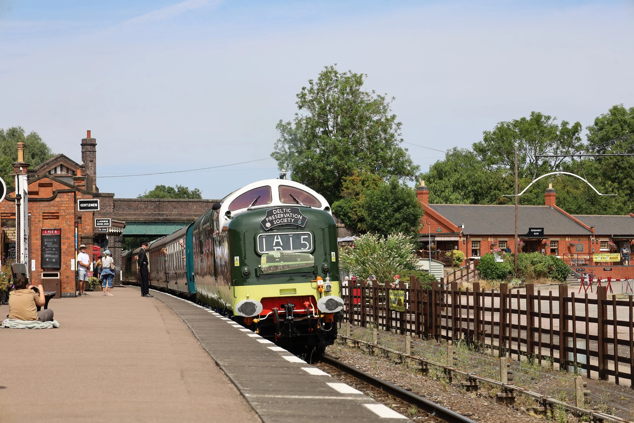 A vintage train station with a green and cream passenger train stopped at the platform, surrounded by people. There are brick buildings, trees, and signs indicating amenities, with a blue sky overhead.
