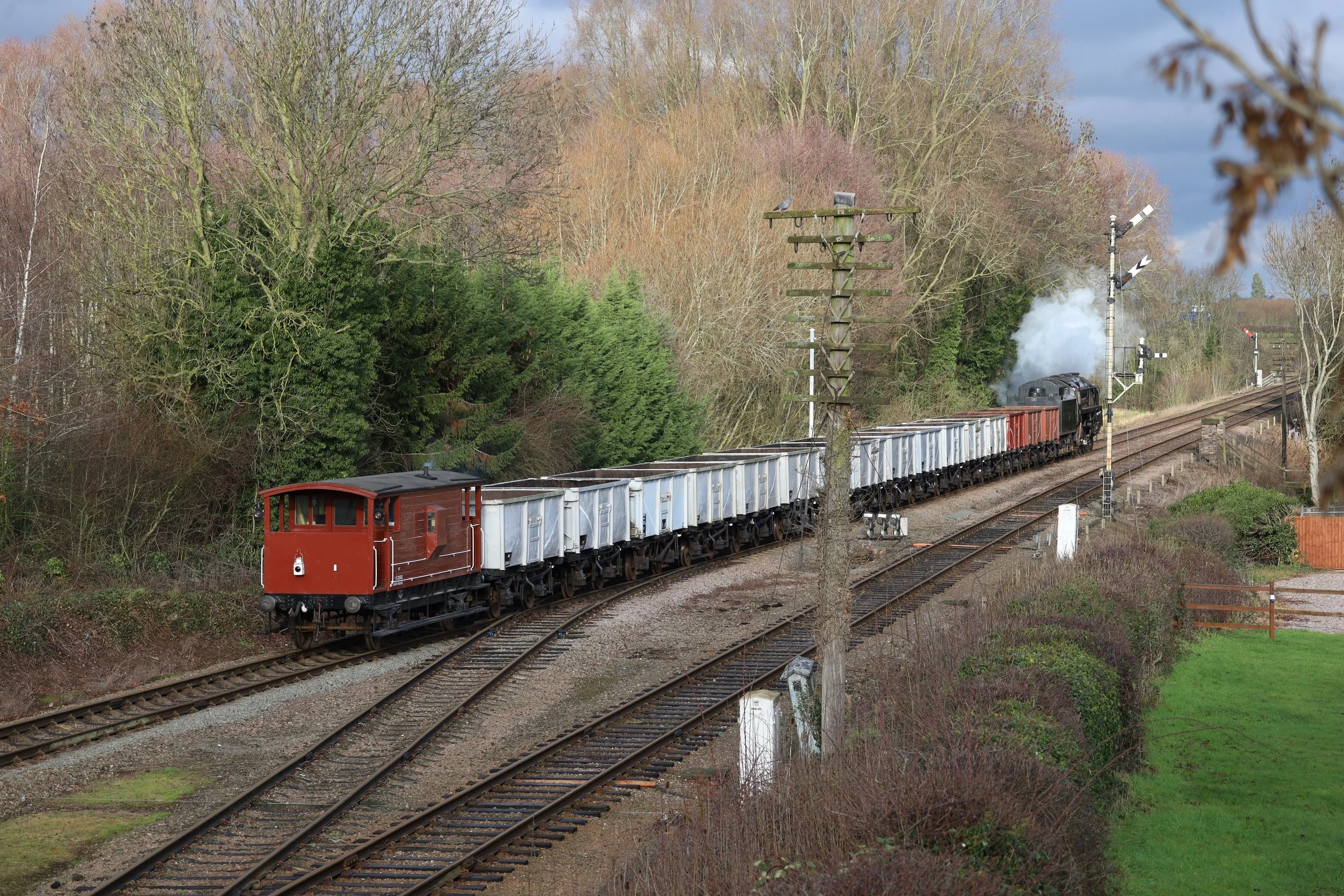 A steam-powered freight train with red locomotive pulling empty freight cars, moving along a curved railway track through a landscape with leafless trees and green bushes, under partly cloudy skies.