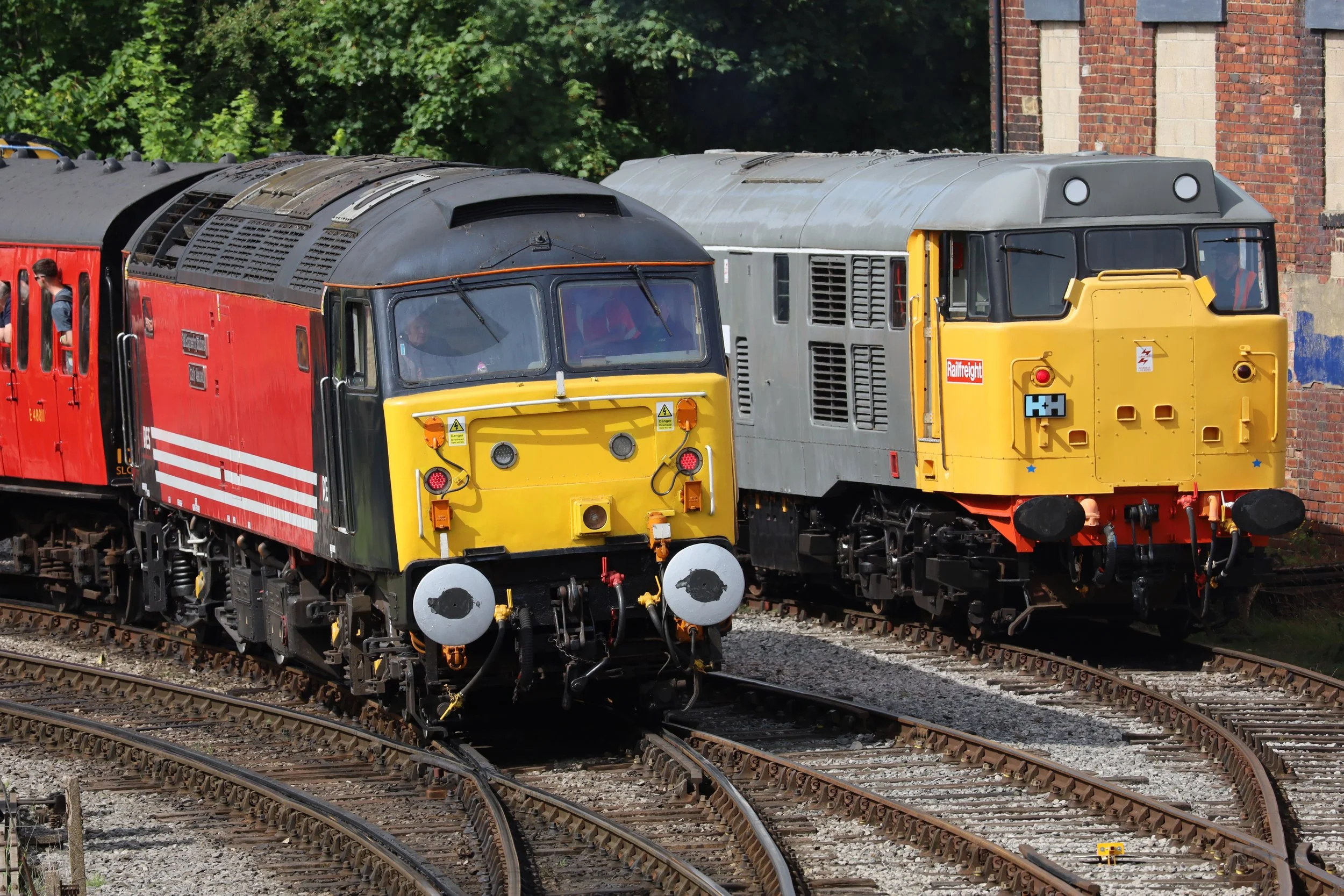 Two colorful train locomotives, one red and one yellow, parked on railway tracks with a brick building and green trees in the background.