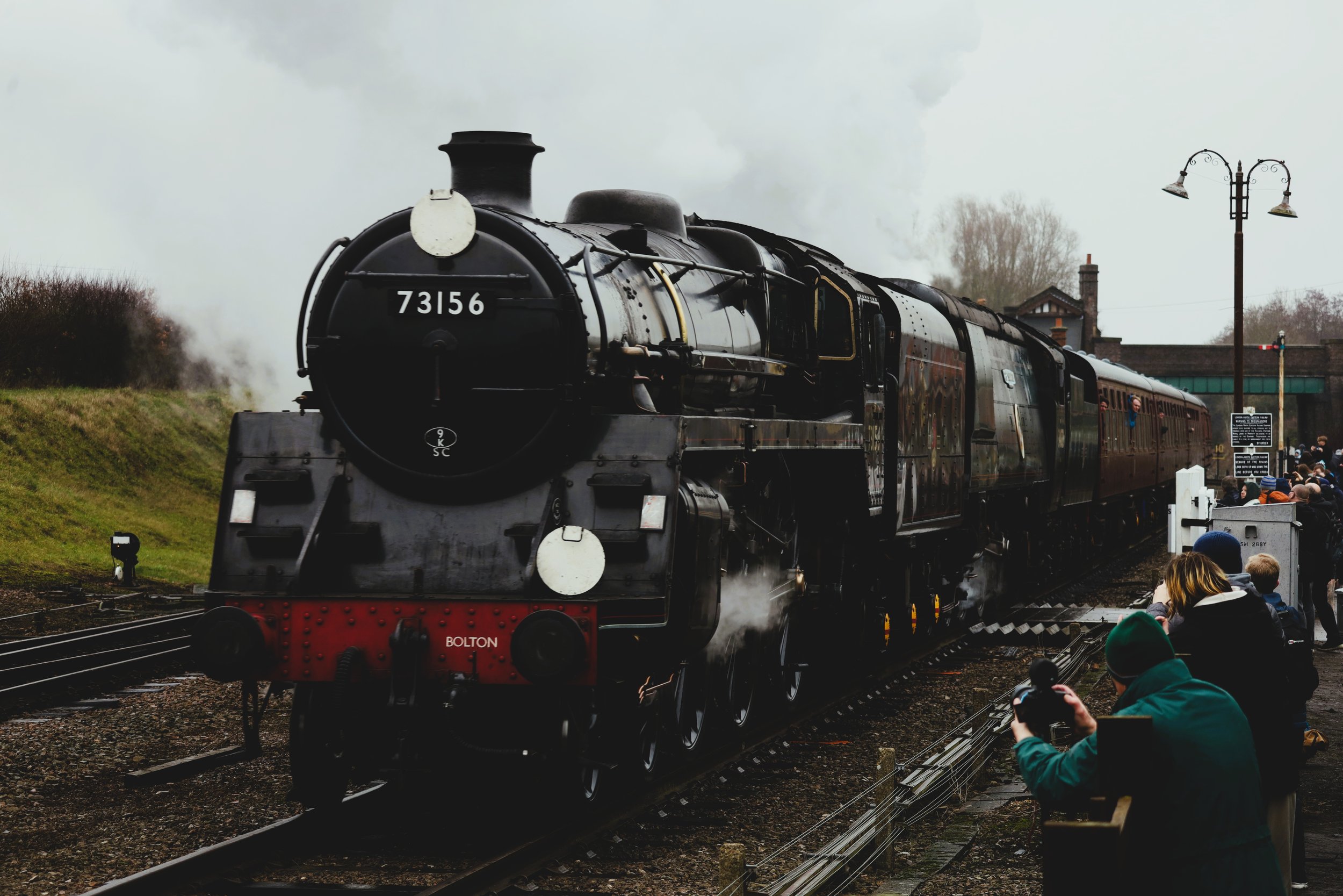A black steam locomotive train labeled 73156 moving on the tracks with people gathered beside the tracks watching and taking photos, in a rural setting with trees and a small building in the background, on an overcast day.