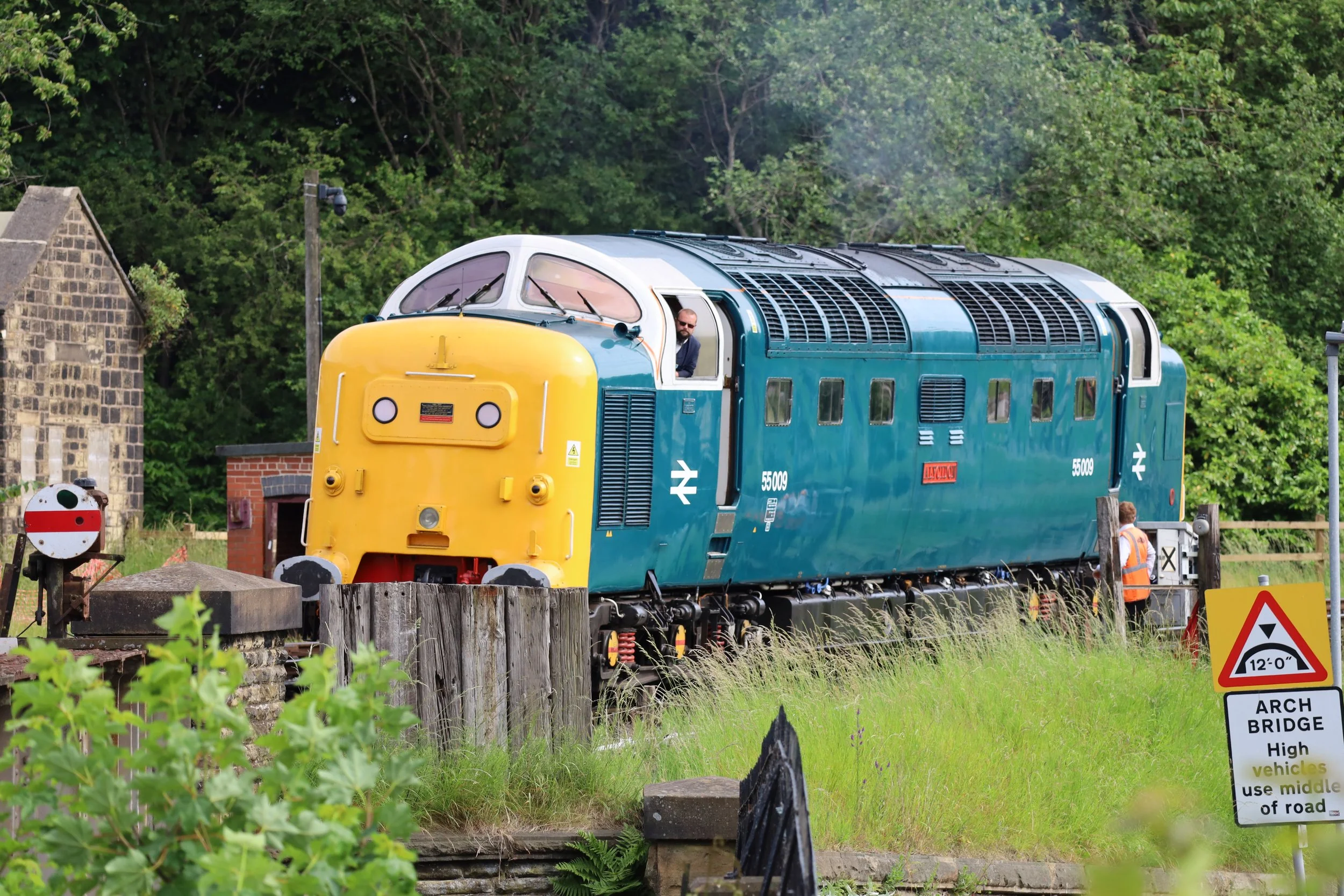 A vintage blue and yellow train moving through a green rural area with trees and buildings in the background. There are signs indicating a high-level bridge and a warning for vehicles to use the middle of the road.