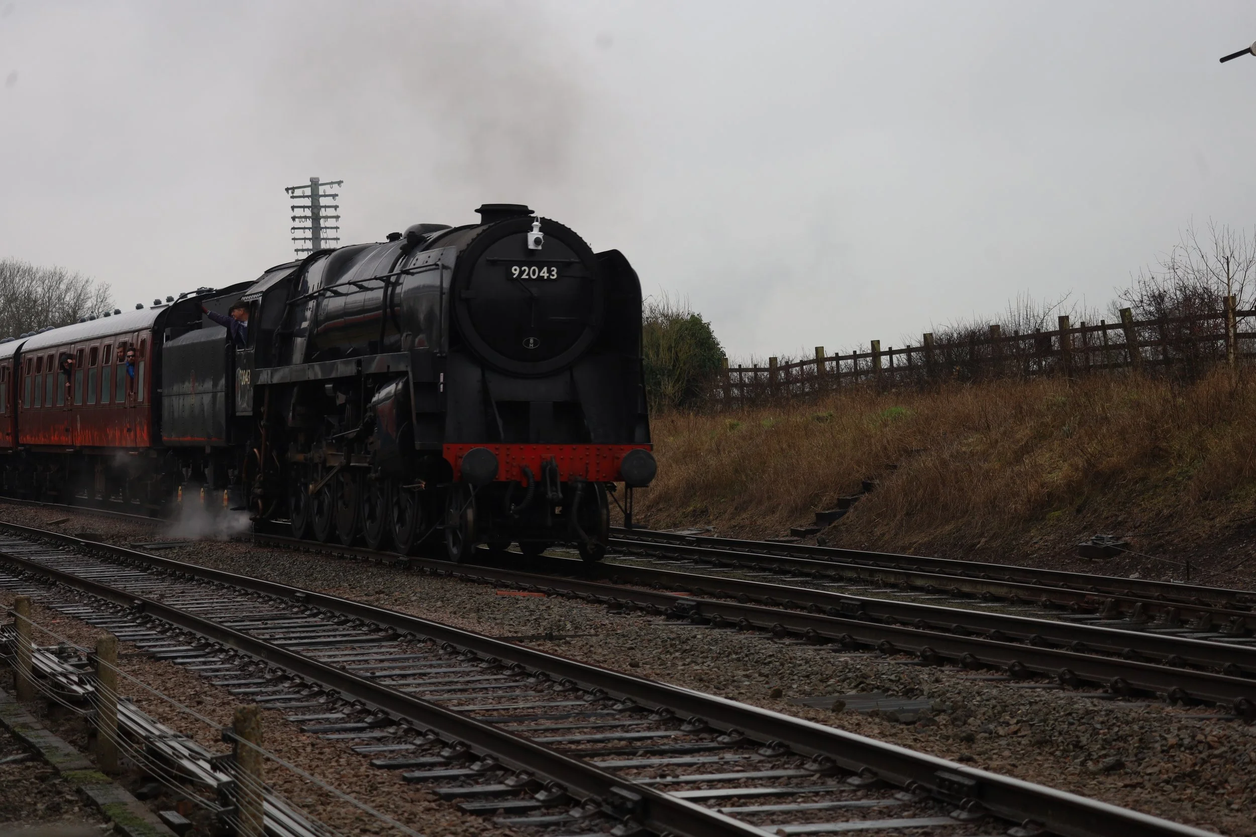 A black steam locomotive train numbered 92043 pulling red passenger cars on railway tracks on a cloudy day with trees and a wooden fence in the background.