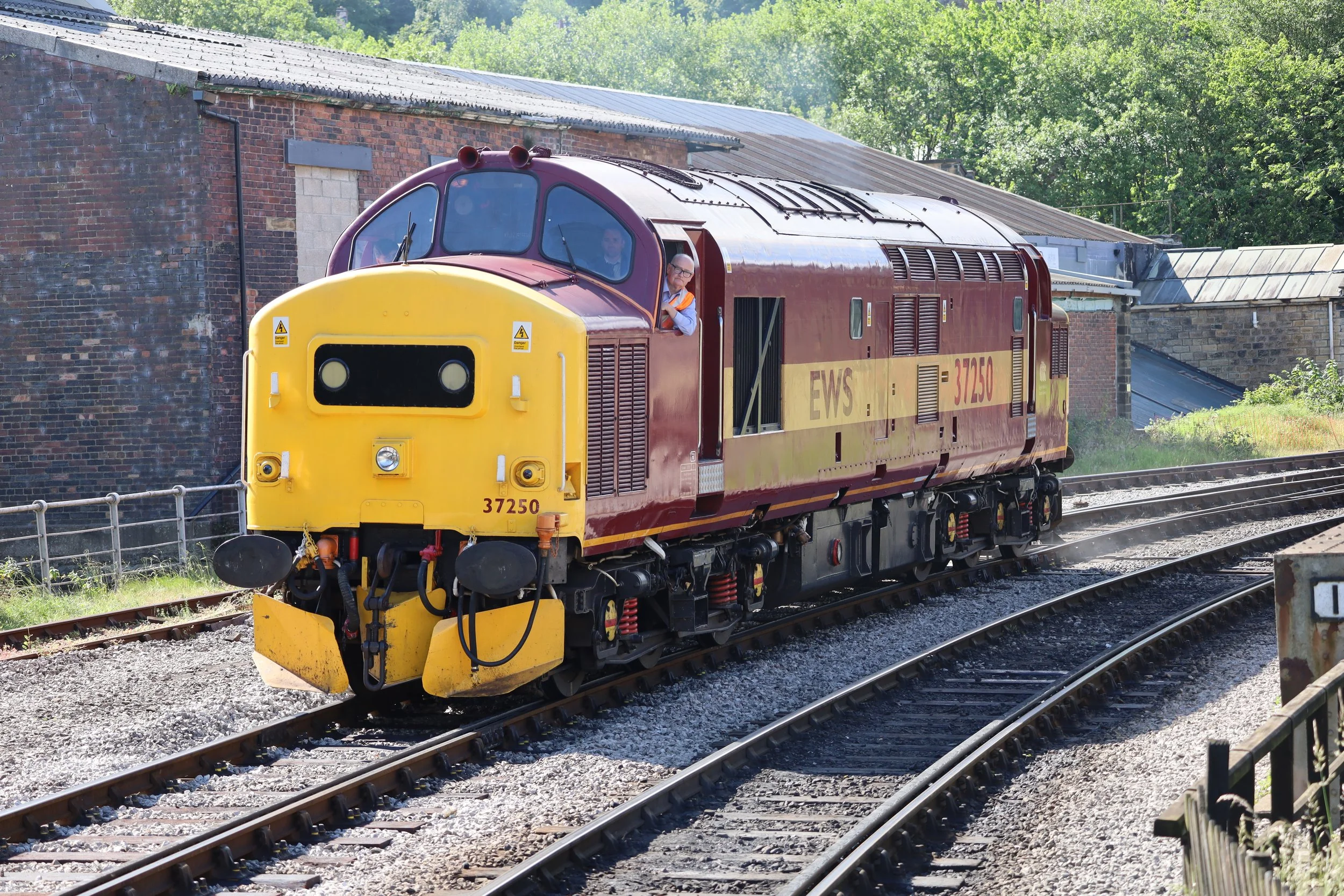 A vintage red and yellow electric locomotive moving along railroad tracks, with a person visible inside the driver's cabin and an industrial brick building and green trees in the background.