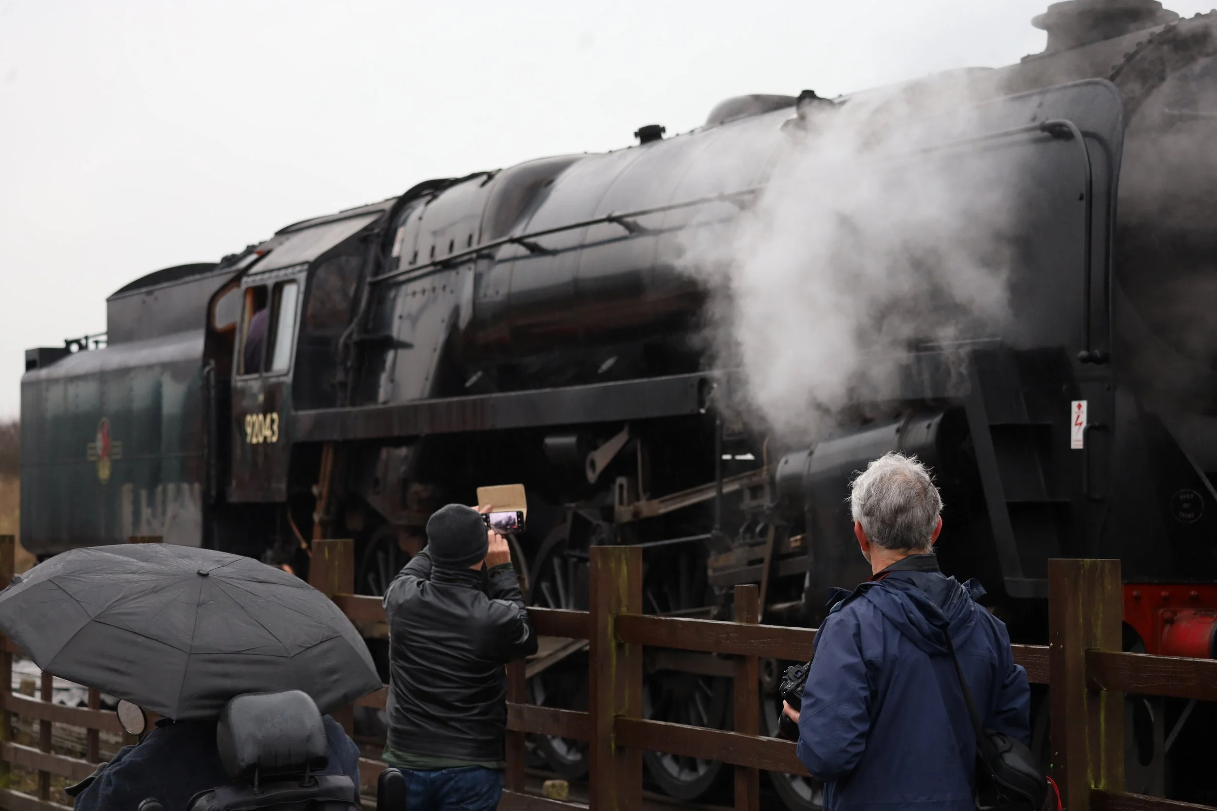 A vintage steam locomotive on display with steam rising from its engine, surrounded by spectators and photographers taking pictures, during an outdoor event on a cloudy day.