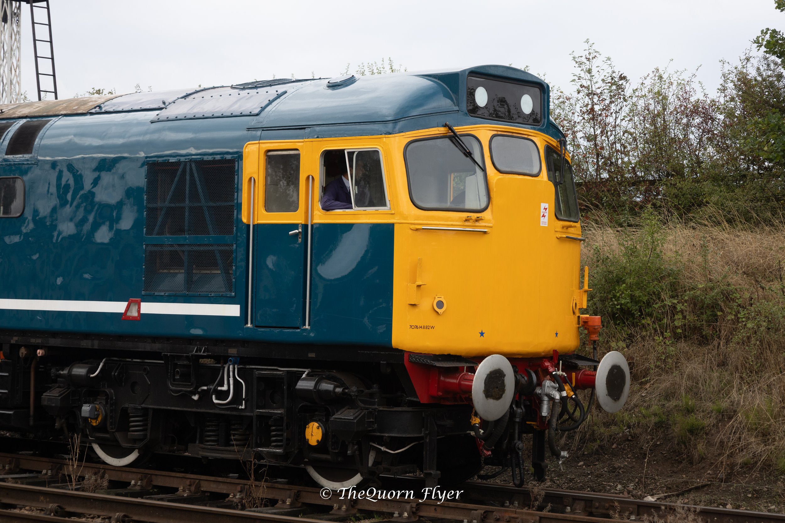 Close-up of a vintage train engine painted in blue and yellow, with a cartoonish face on the front, traveling on railroad tracks and surrounded by greenery.