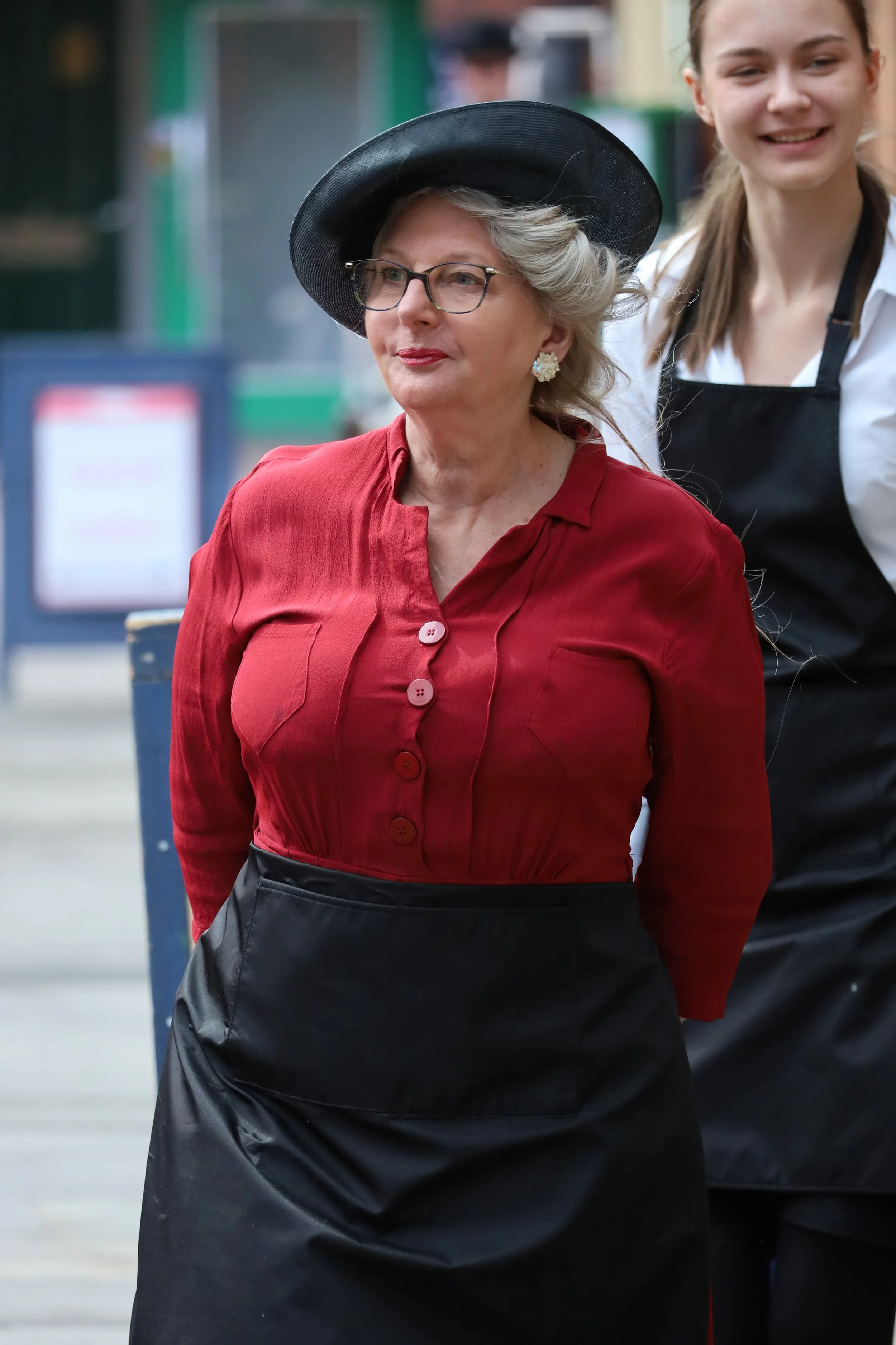 A woman with glasses and pearl earrings wearing a black hat and a red blouse standing next to a smiling younger woman with a white shirt and apron.
