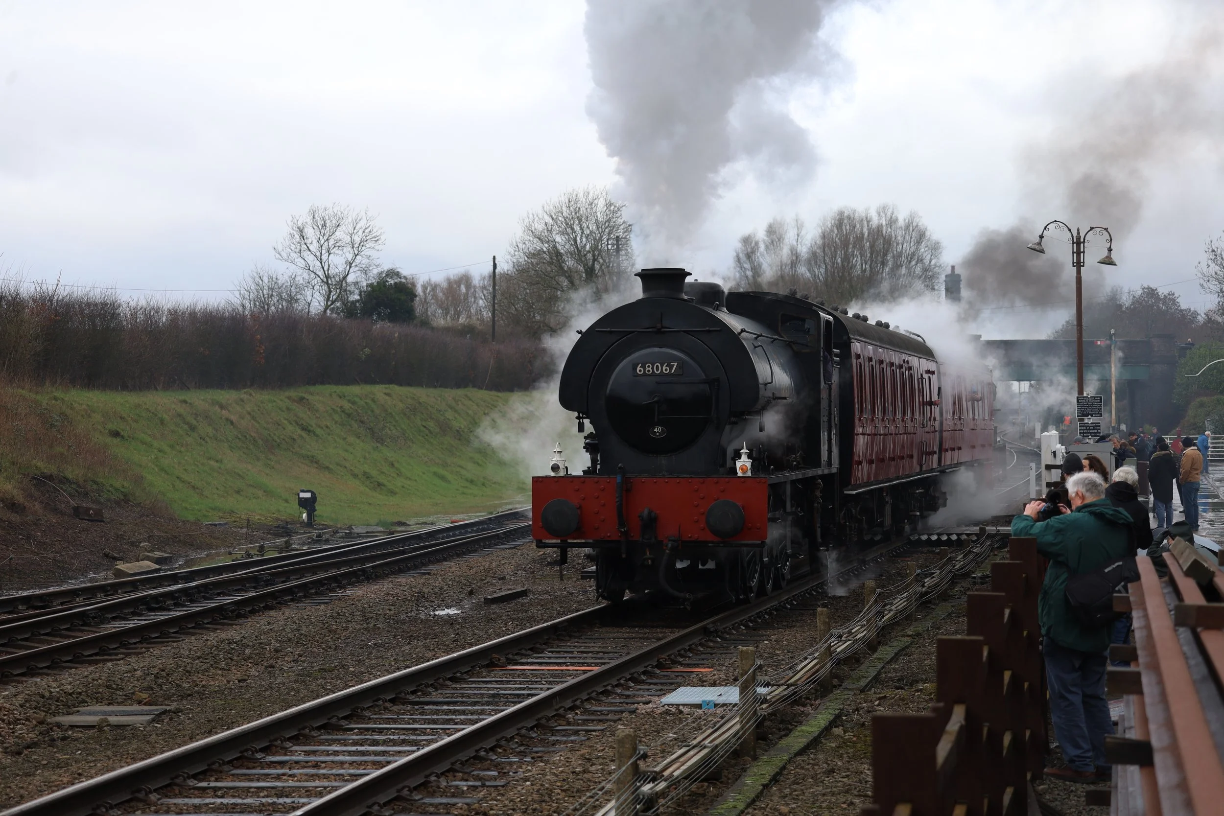 A vintage steam train moving on railway tracks, emitting black smoke, with onlookers and photographers on the platform.
