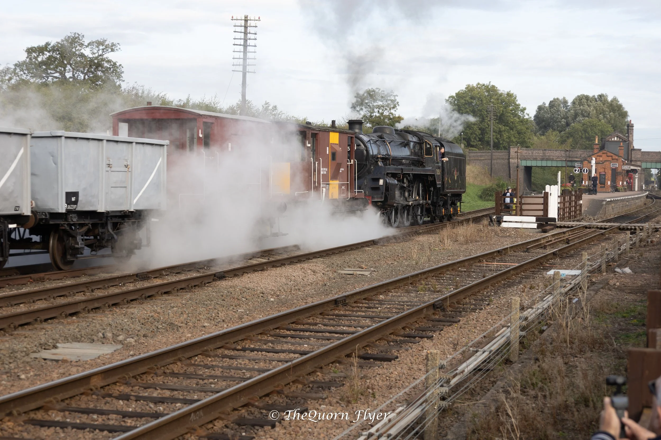 A vintage steam train moving along railway tracks with smoke coming out of its chimney, at a historic train station with some people waiting on the platform and trees in the background.