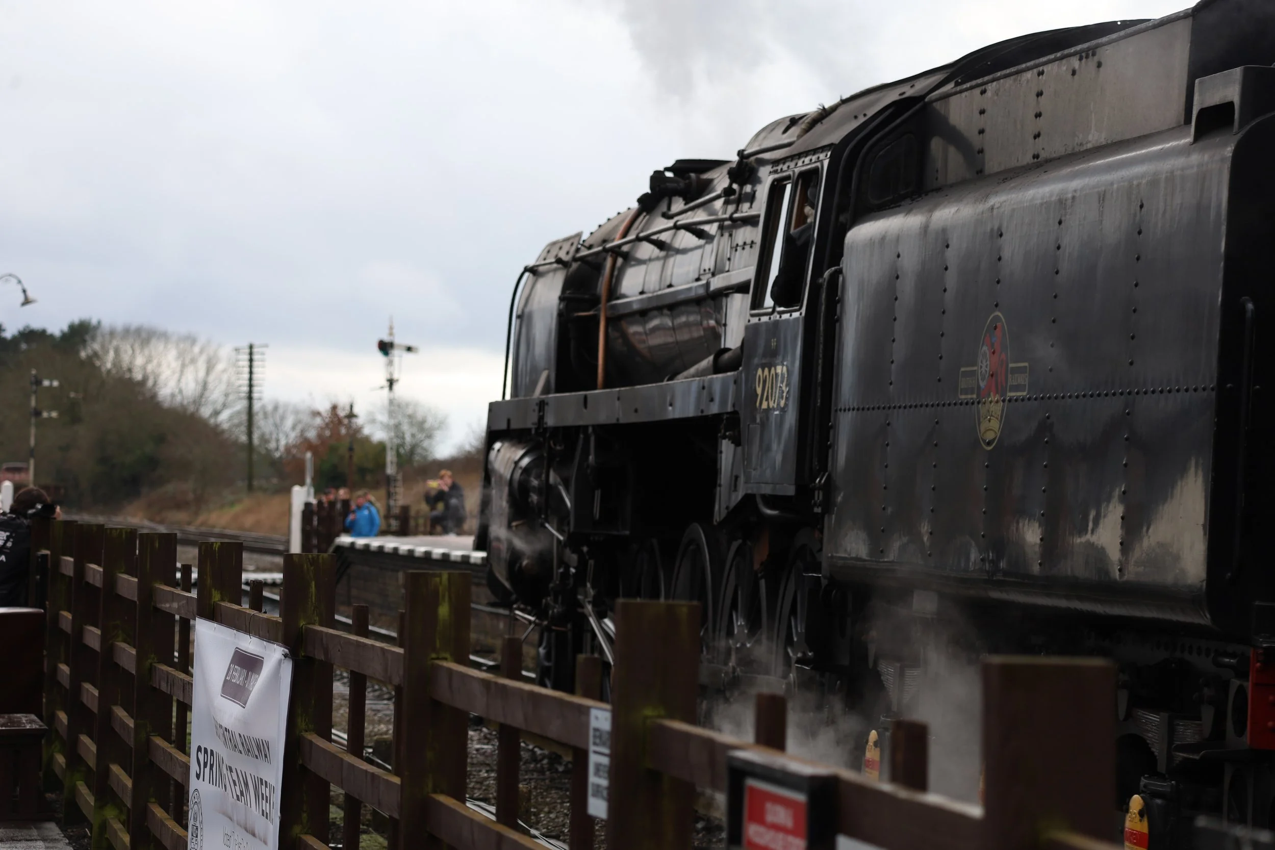 Close-up of a vintage black steam locomotive at a train station, with part of a crowd visible in the background.