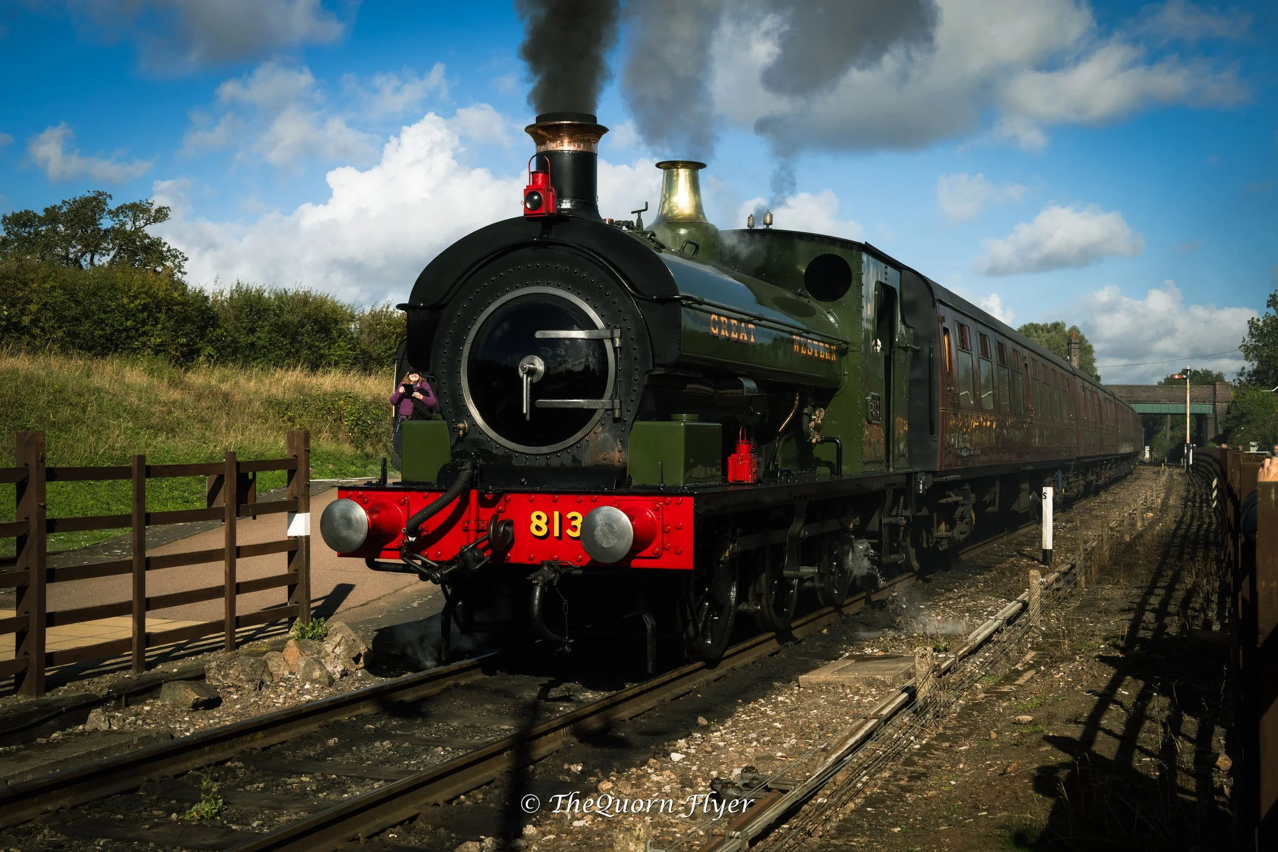 A vintage black and green steam train with the number 813 on the front, pulling into a station with smoke billowing from its chimney. There is a person in a purple jacket taking a photo near the train. The sky is partly cloudy with patches of blue, and there is greenery in the background.