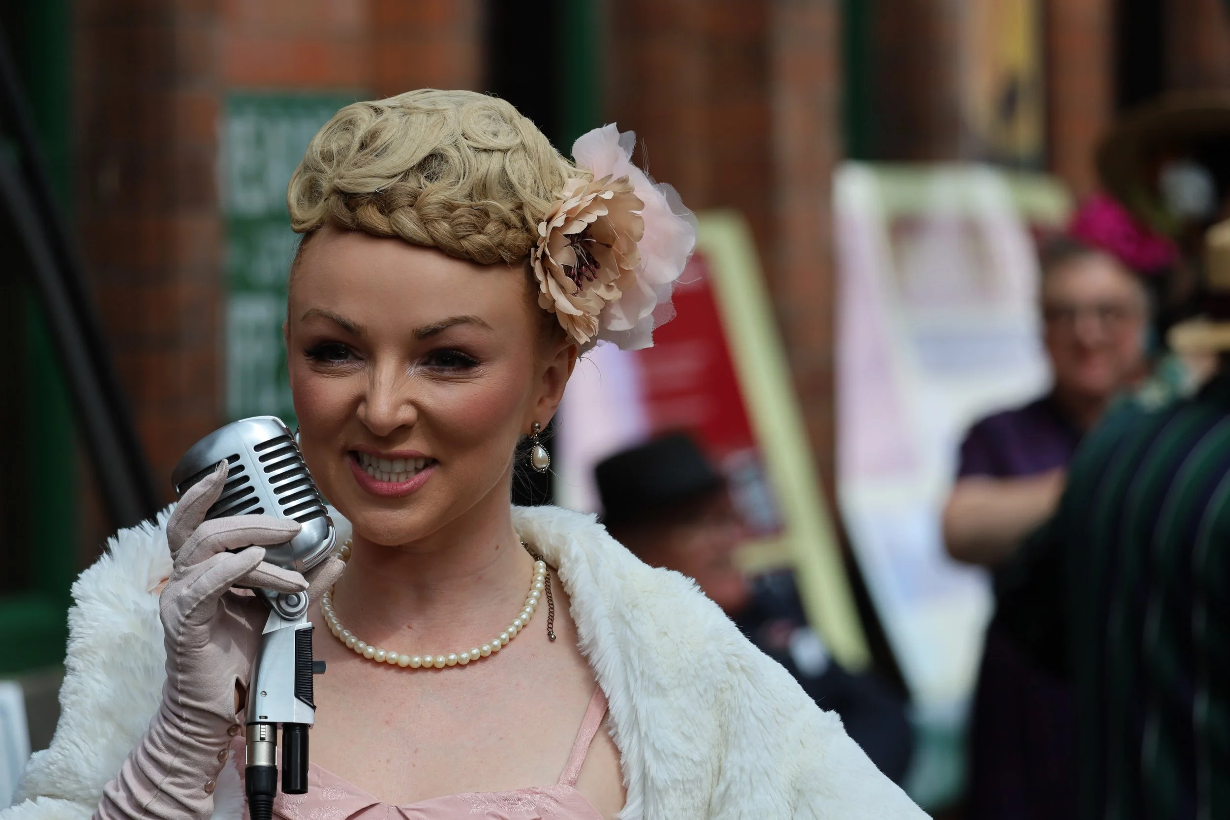 A woman dressed in vintage attire, wearing a pearl necklace and gloves, holding a microphone, with a decorative flower in her hair, appearing to be speaking or singing at an event.