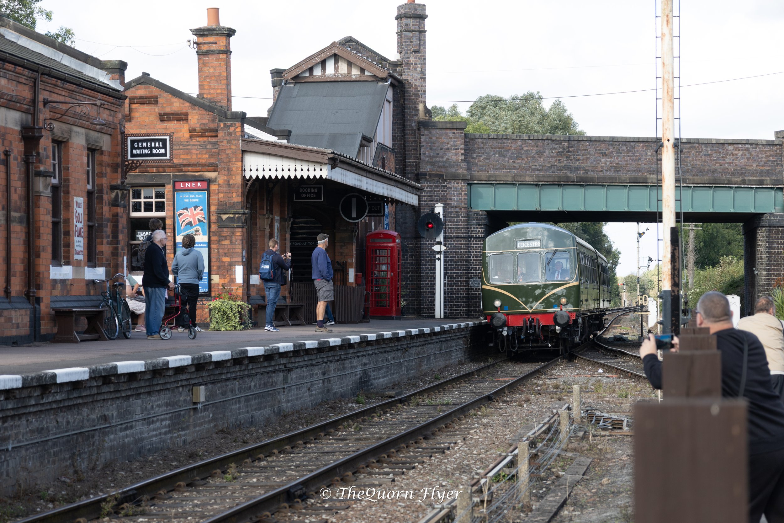 A vintage train arriving at a historic station with people waiting on the platform. The station building is made of red brick, with a sign reading 'General Waiting Room' and a red British telephone booth nearby. The scene includes a green train, a bridge overhead, and several individuals, some taking photos and others standing or sitting on benches.