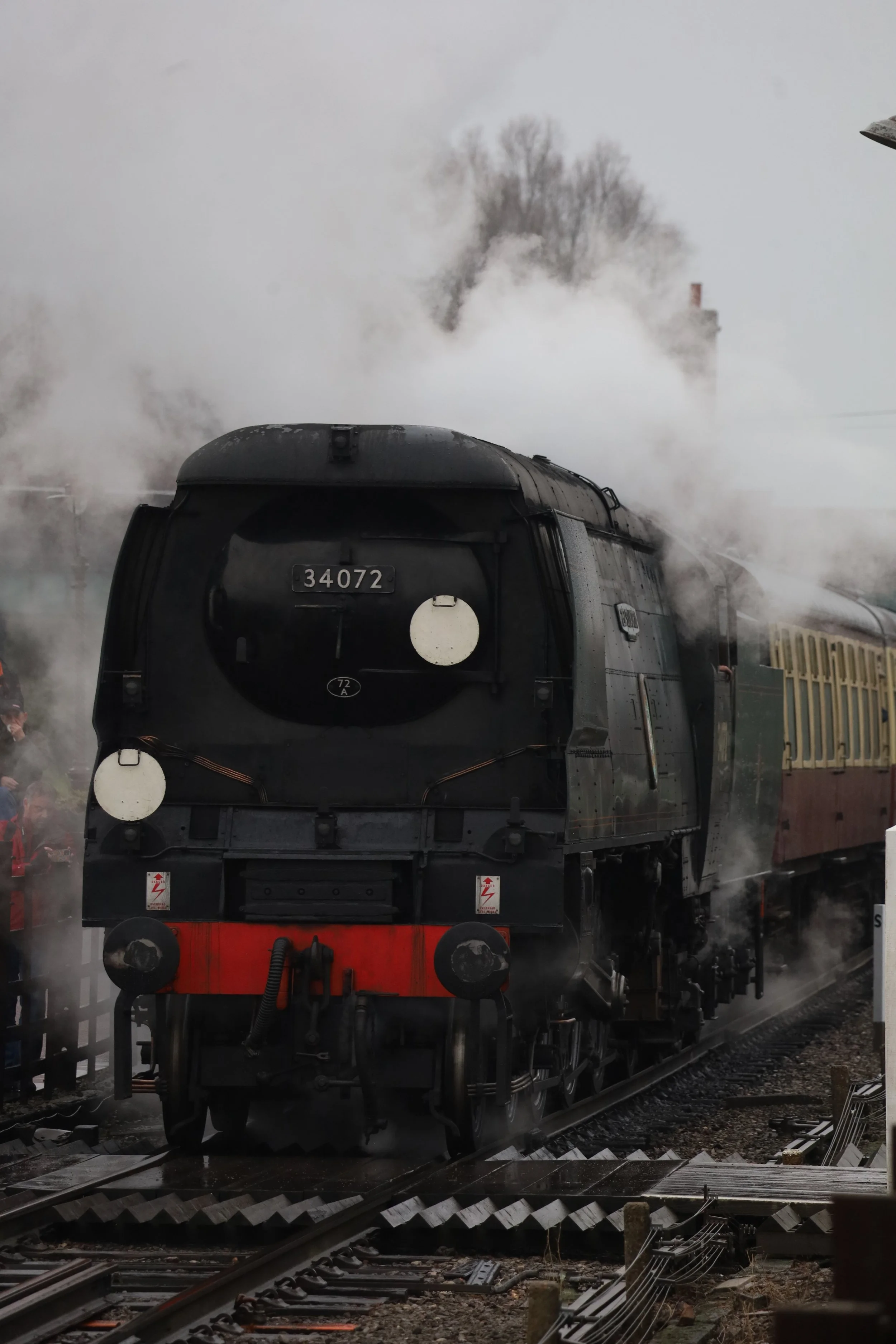 A black steam locomotive train with the number 34072 on its front, emitting steam and smoke, on railway tracks with station platform and trees in the background.